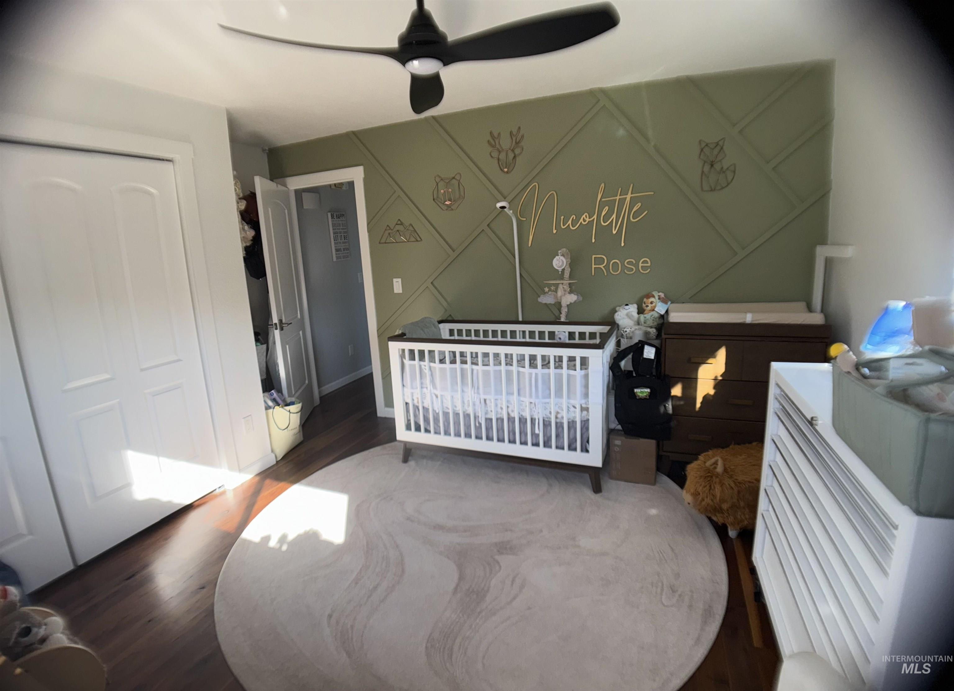 Bedroom with a nursery area, wood finished floors, a ceiling fan, a closet, and a decorative wall