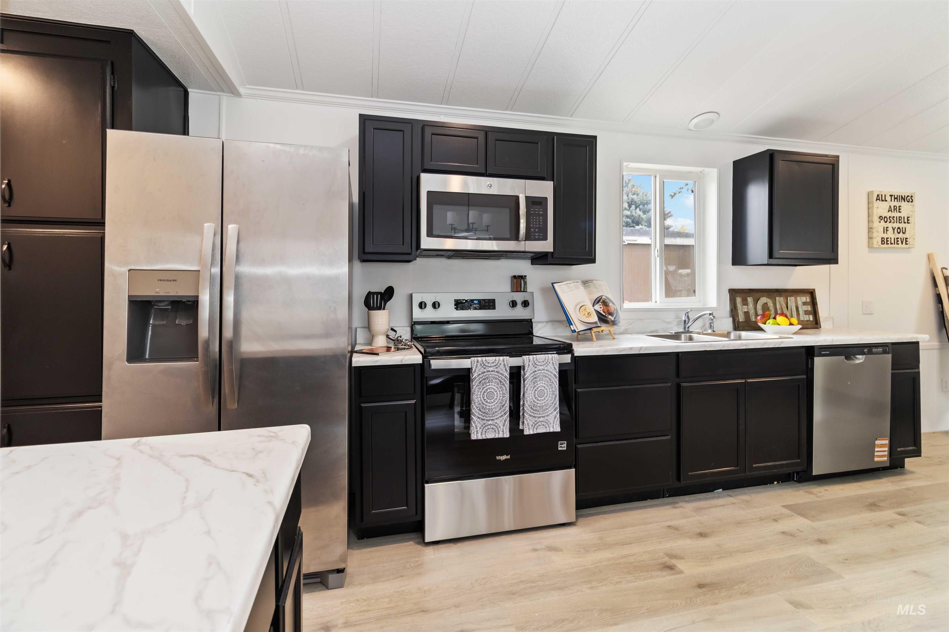 Kitchen with dark cabinets, stainless steel appliances, light wood-style flooring, light stone counters, and crown molding