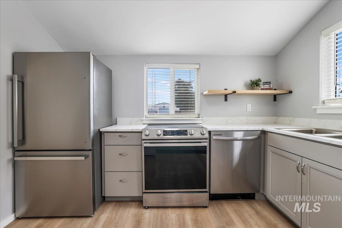 Kitchen featuring appliances with stainless steel finishes, light wood finished floors, gray cabinetry, open shelves, and light stone counters