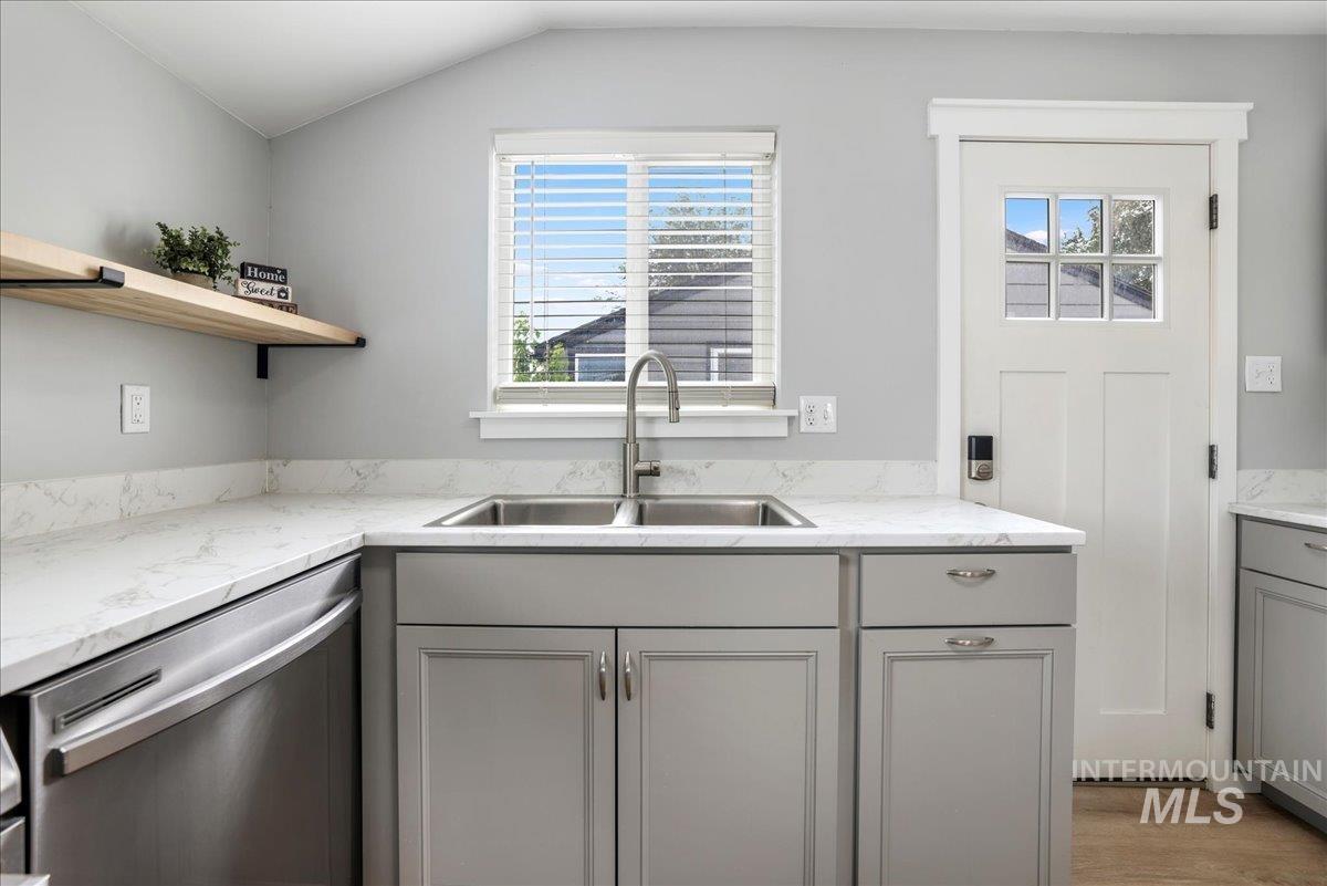 Kitchen with gray cabinetry, stainless steel dishwasher, light stone counters, and vaulted ceiling