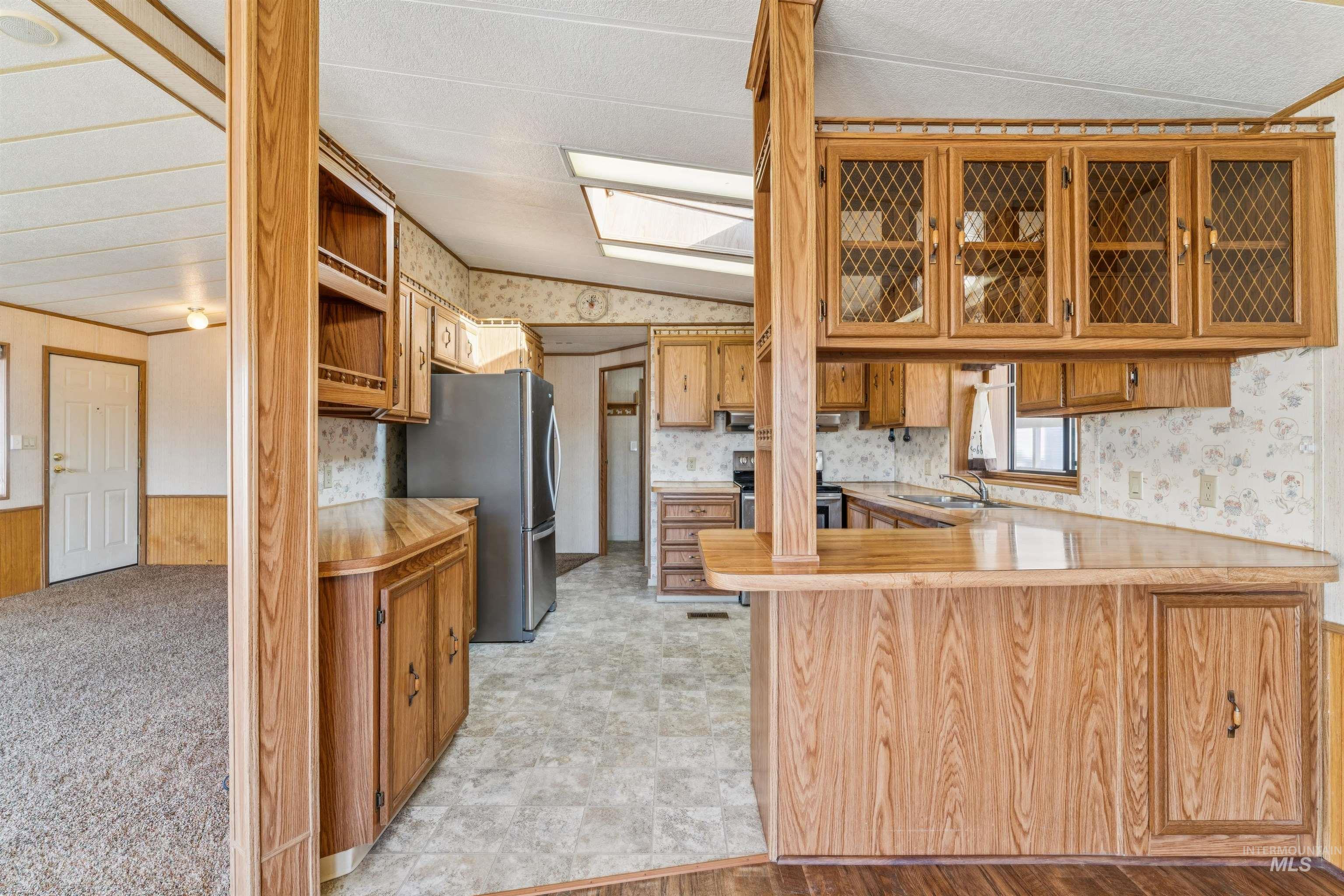 Kitchen with wallpapered walls, a peninsula, a wainscoted wall, light countertops, and glass insert cabinets