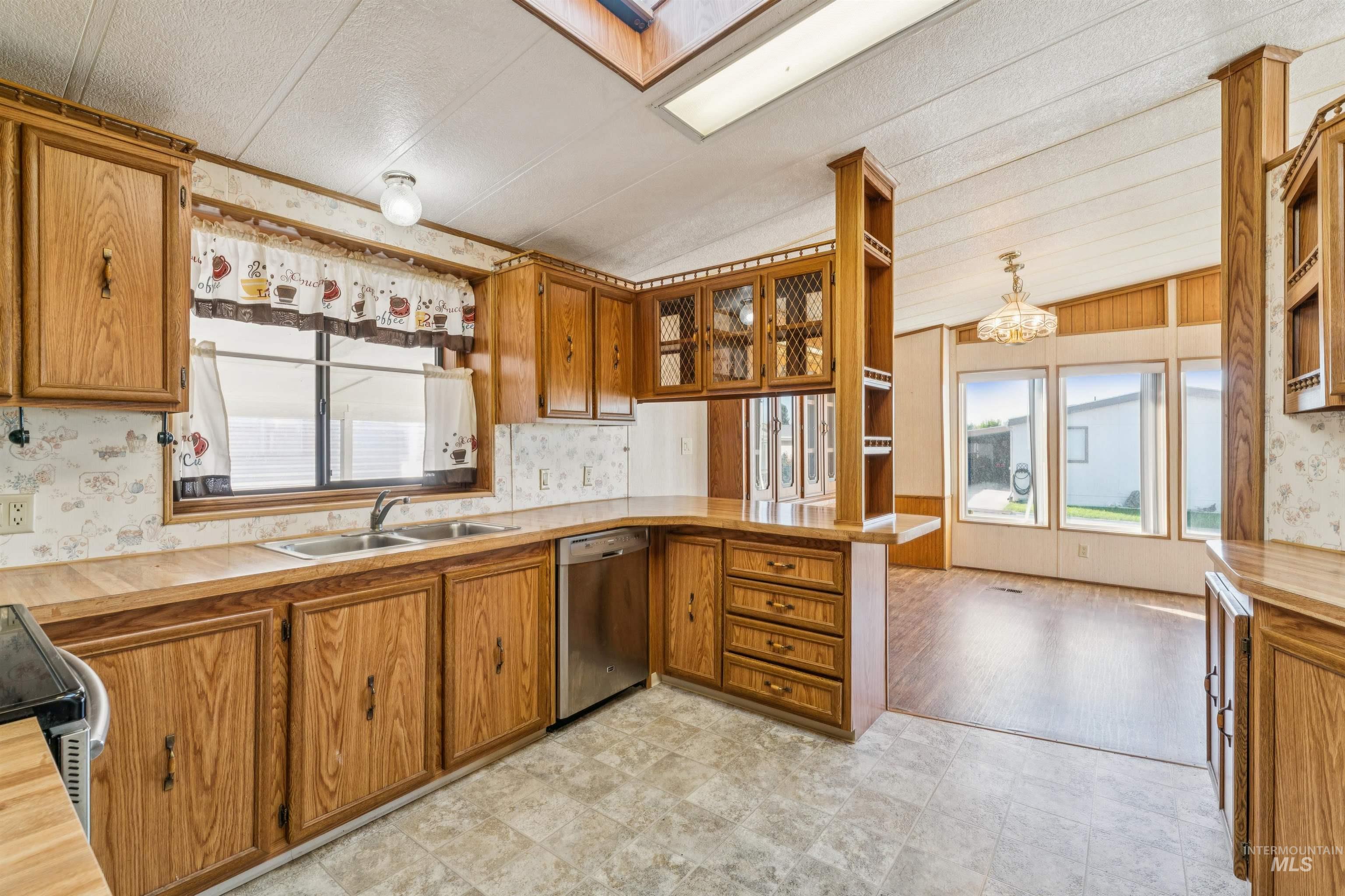 Kitchen featuring wallpapered walls, a peninsula, brown cabinets, light countertops, and stainless steel dishwasher