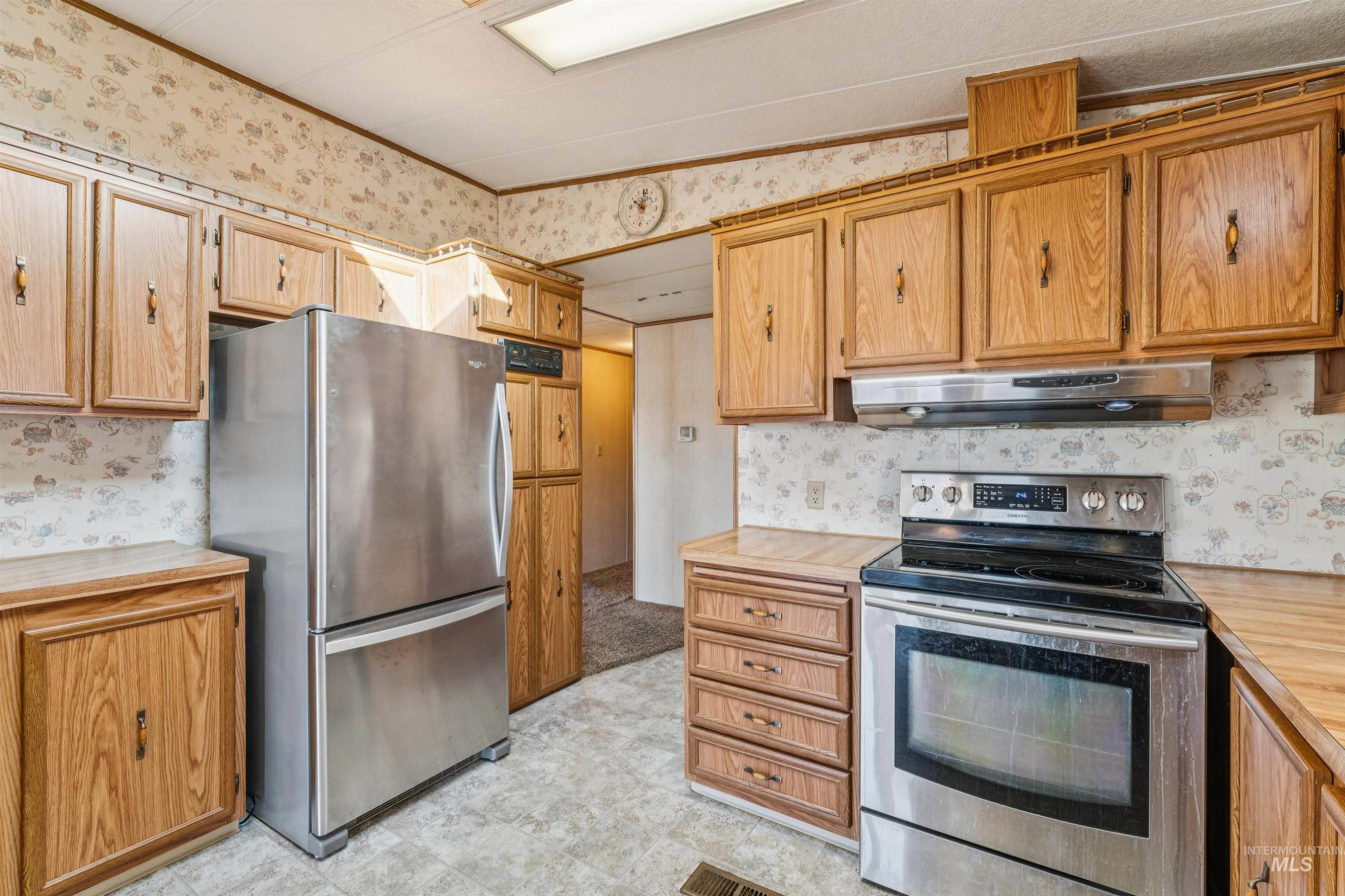 Kitchen featuring wallpapered walls, appliances with stainless steel finishes, under cabinet range hood, butcher block counters, and brown cabinets