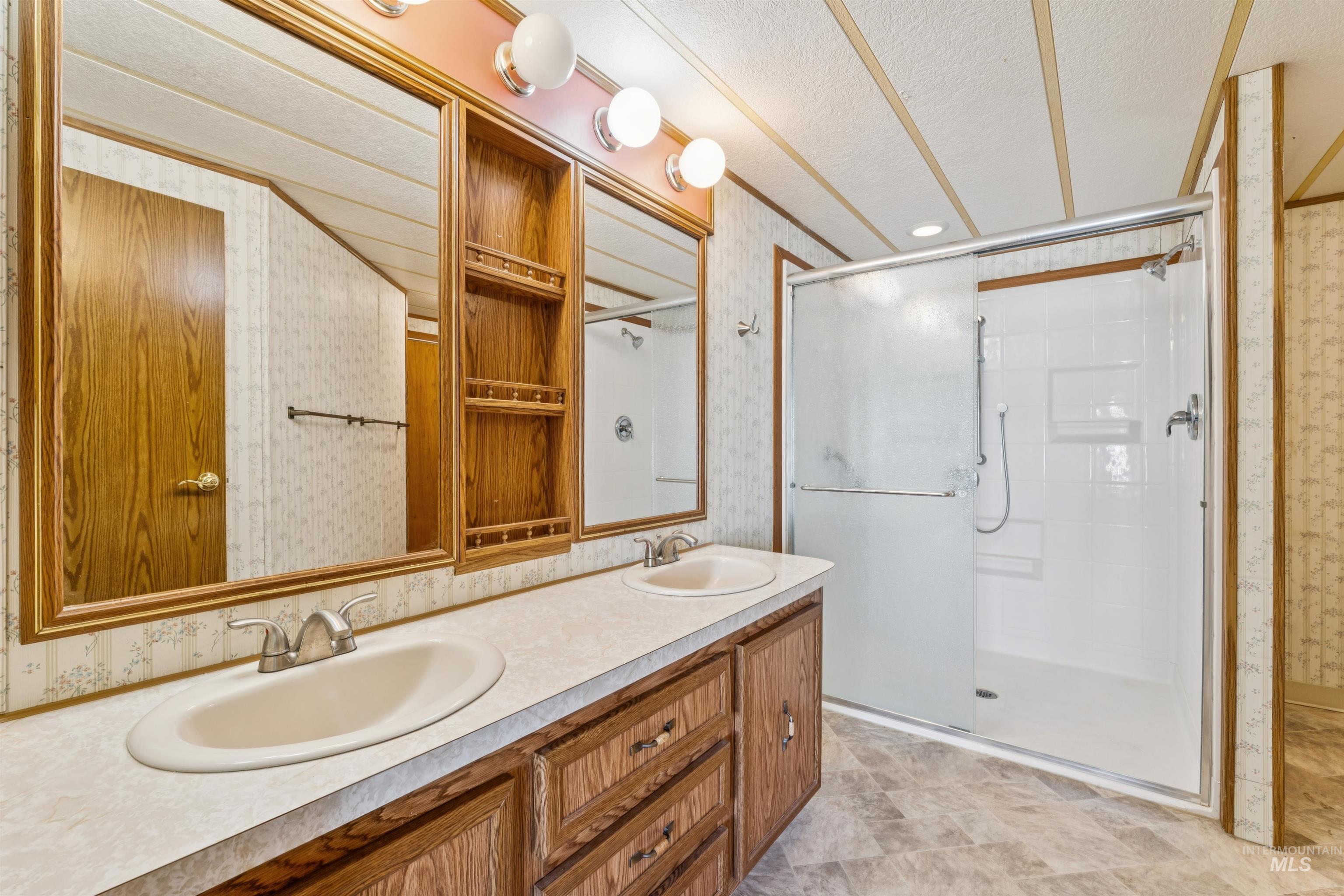 Bathroom featuring wallpapered walls, a textured ceiling, a shower stall, and double vanity