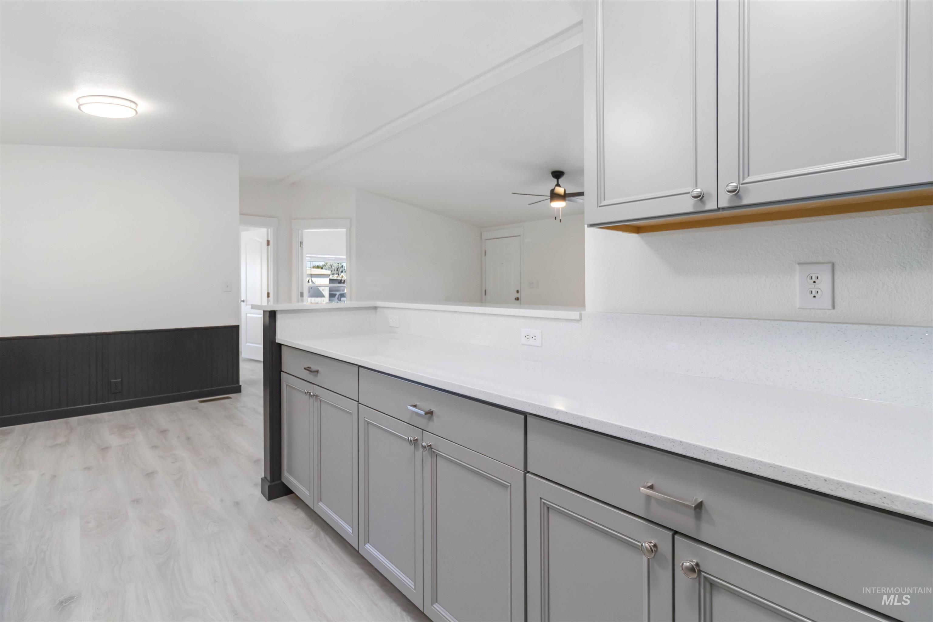 Kitchen featuring gray cabinetry, wainscoting, light wood finished floors, ceiling fan, and light stone counters