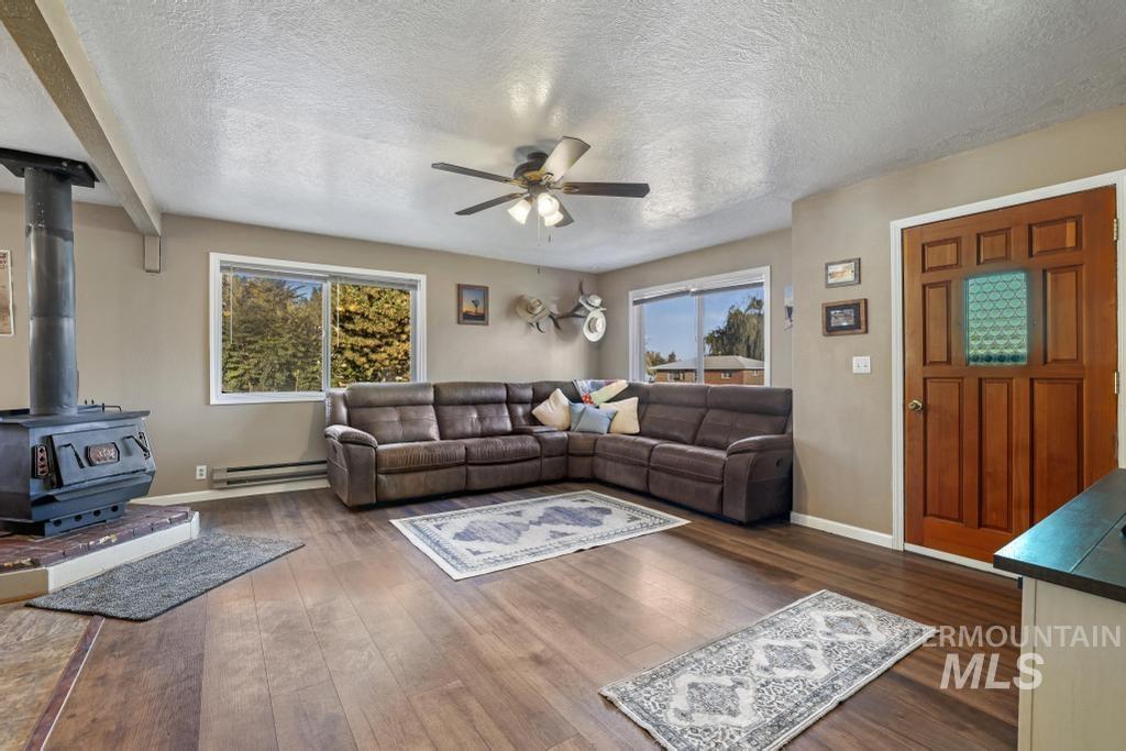 Living room with a wood stove, a textured ceiling, dark wood-type flooring, a ceiling fan, and a baseboard heating unit