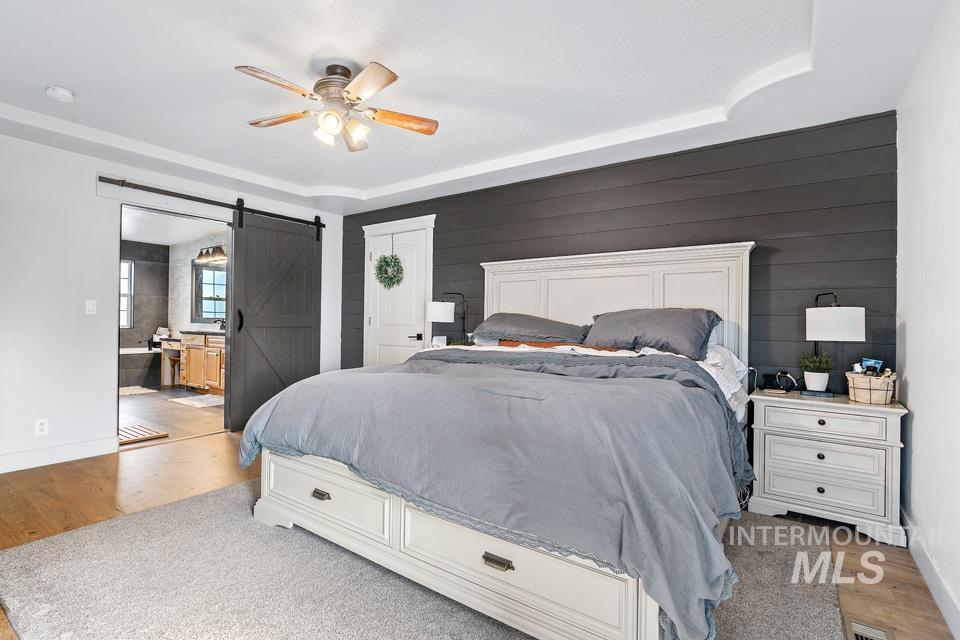 Bedroom featuring a barn door, light wood-type flooring, a ceiling fan, wood walls, and ensuite bathroom