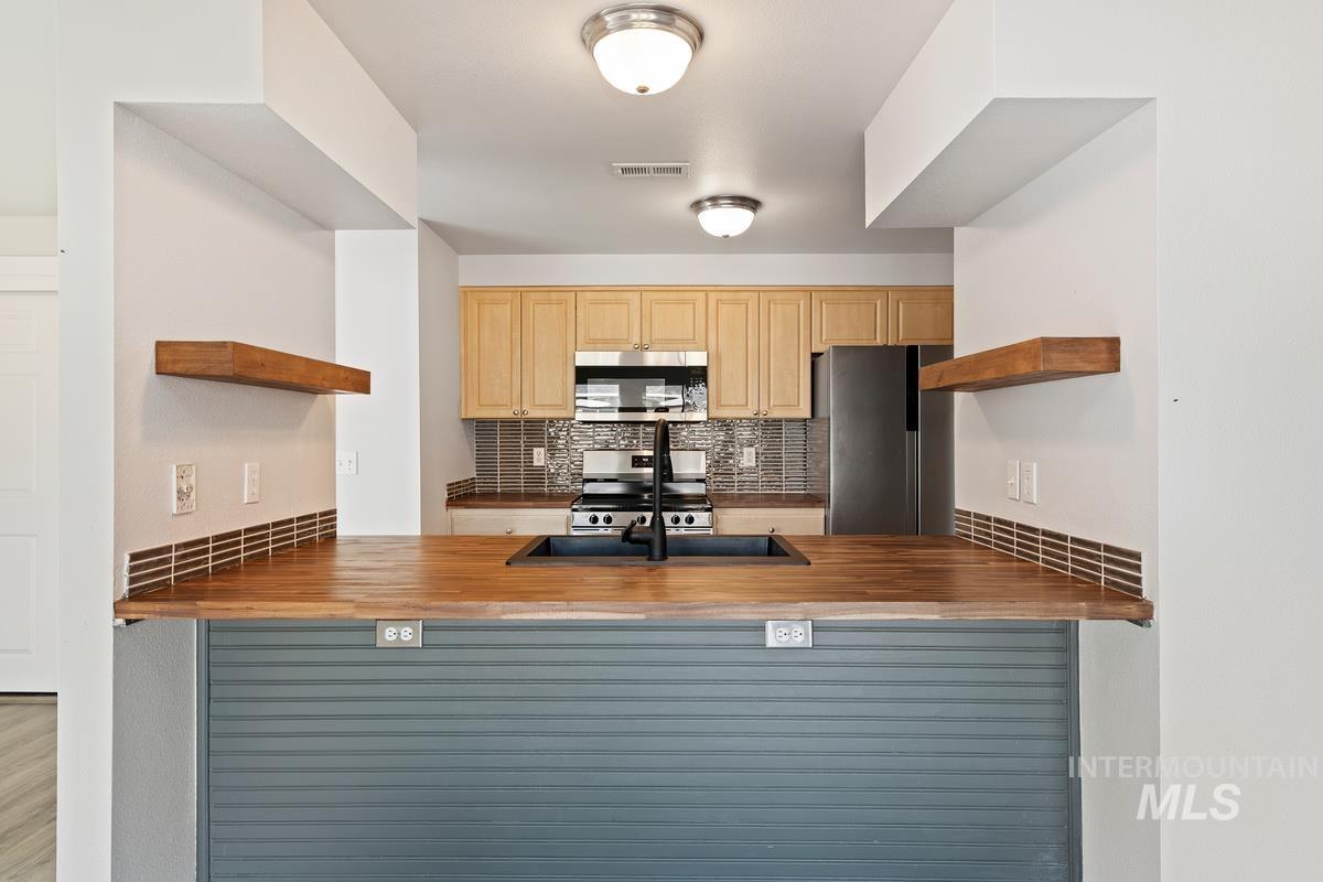 Kitchen with butcher block counters, open shelves, decorative backsplash, stainless steel appliances, and light brown cabinetry