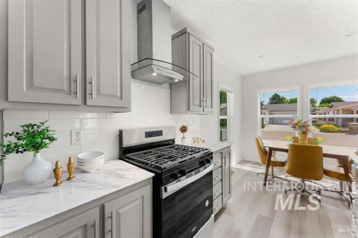 Kitchen with gas stove, wall chimney exhaust hood, backsplash, light wood-style flooring, and light stone countertops