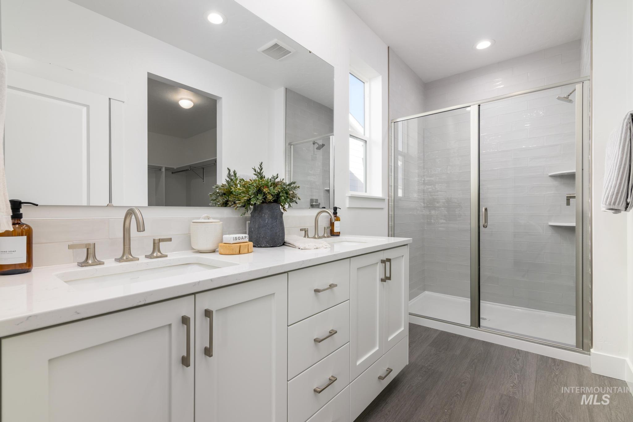 Full bath with dark wood finished floors, a shower stall, double vanity, a spacious closet, and recessed lighting