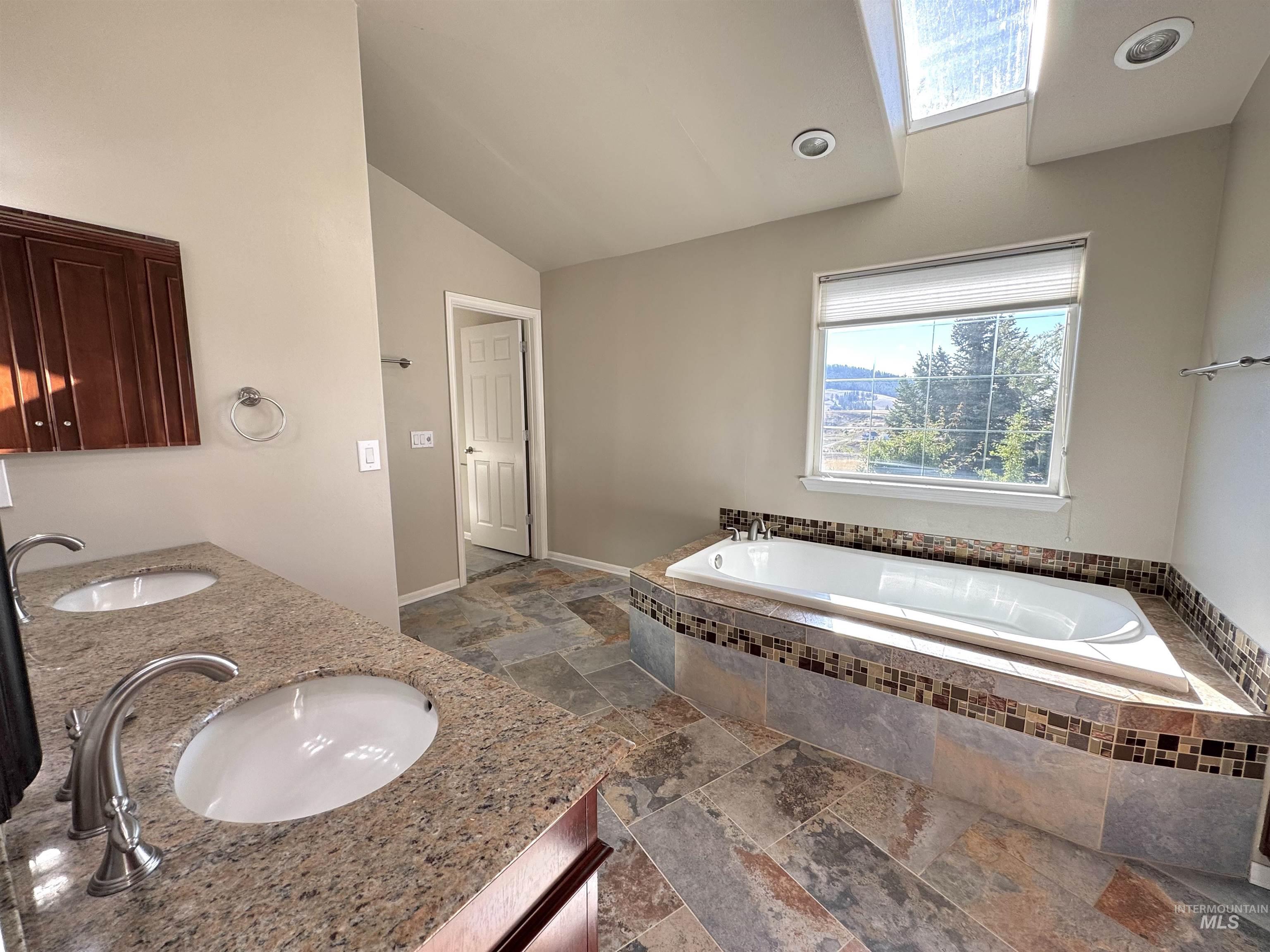 Full bathroom with a garden tub, stone tile flooring, vaulted ceiling, and double vanity