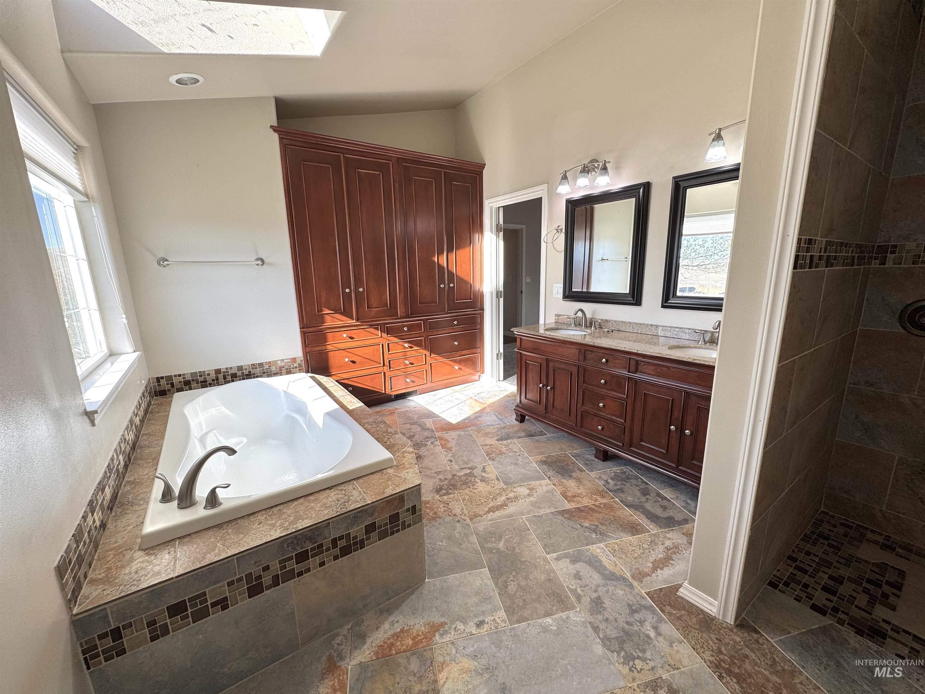 Bathroom featuring double vanity, a bath, walk in shower, lofted ceiling, and stone tile floors