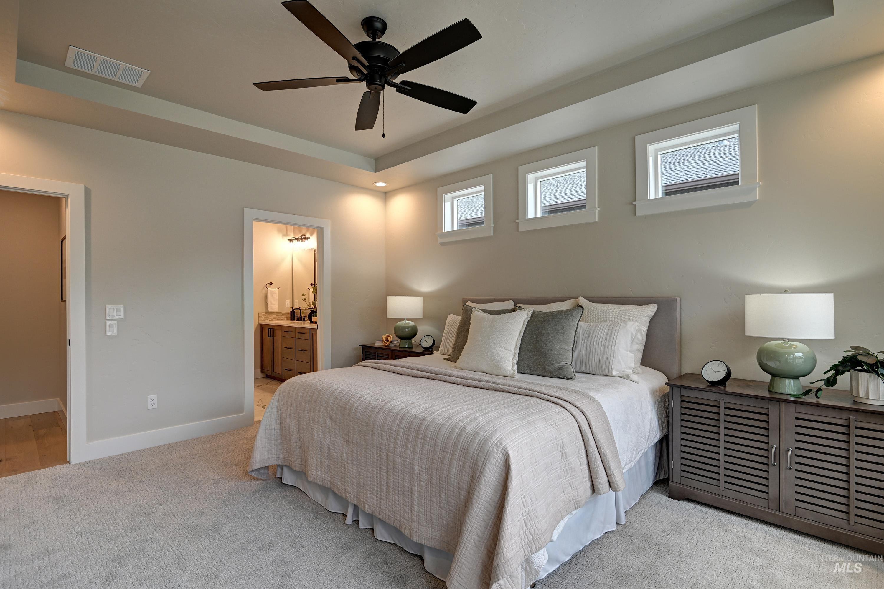 Bedroom with a raised ceiling, light carpet, ensuite bath, and a ceiling fan