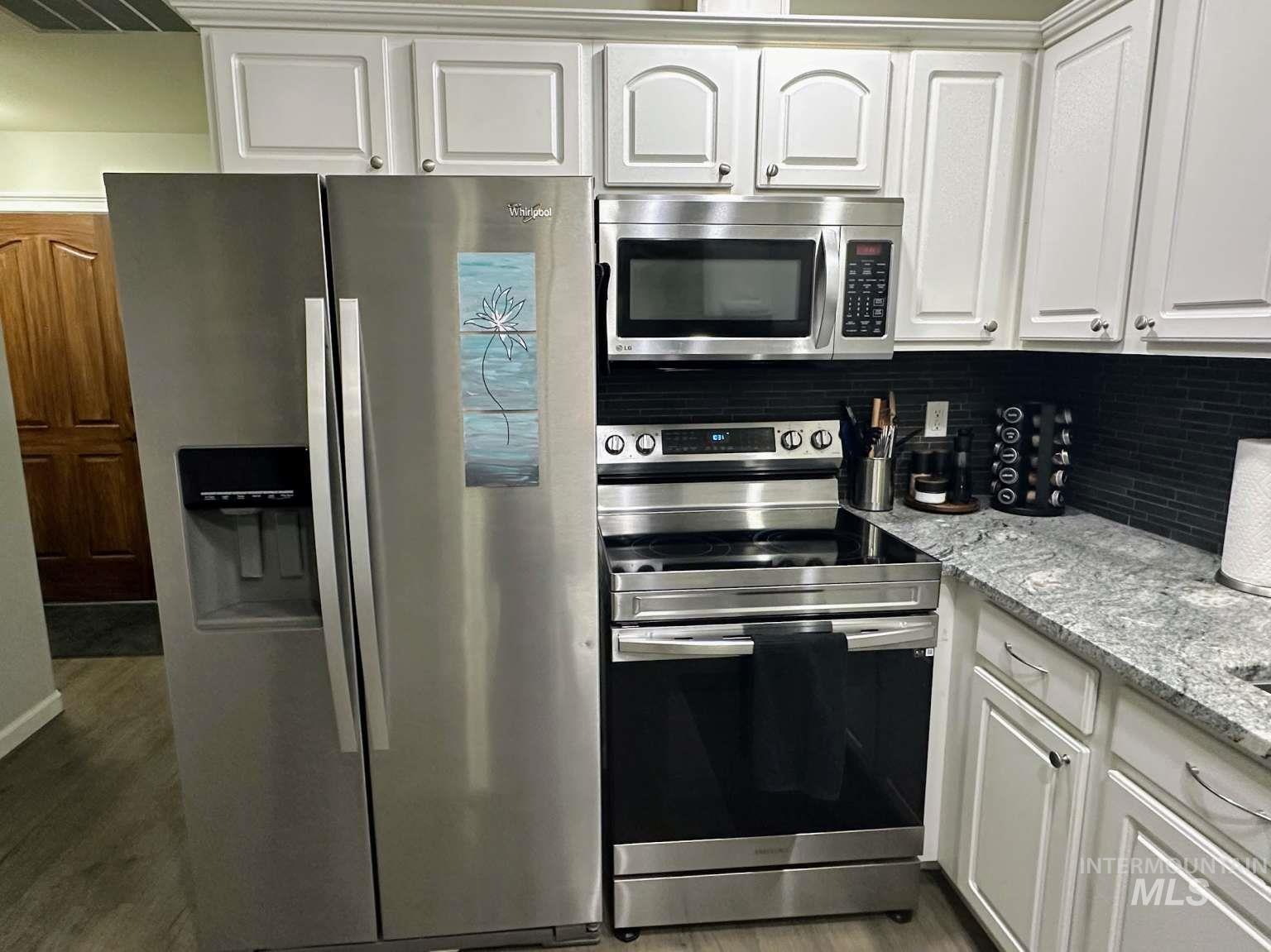 Kitchen with stainless steel appliances, white cabinets, light stone countertops, dark wood-type flooring, and tasteful backsplash