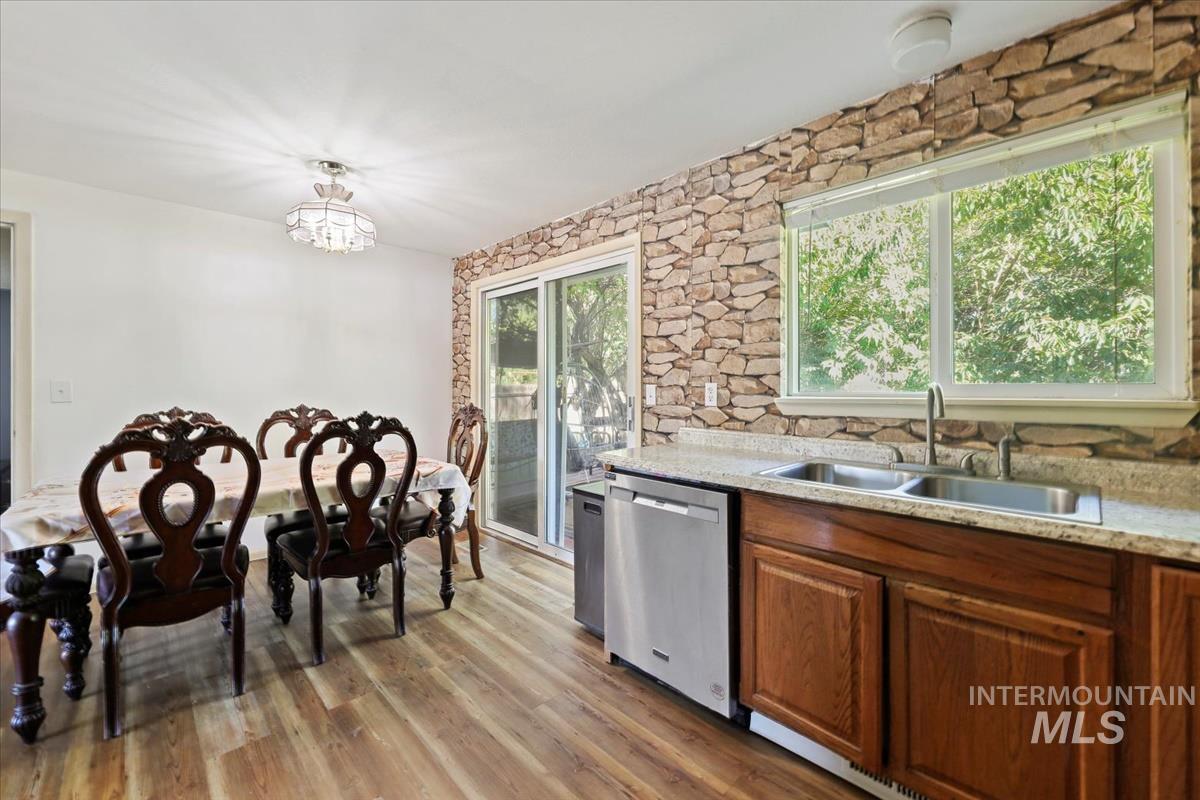 Kitchen with stainless steel dishwasher, light wood-type flooring, hanging light fixtures, brown cabinets, and light stone counters