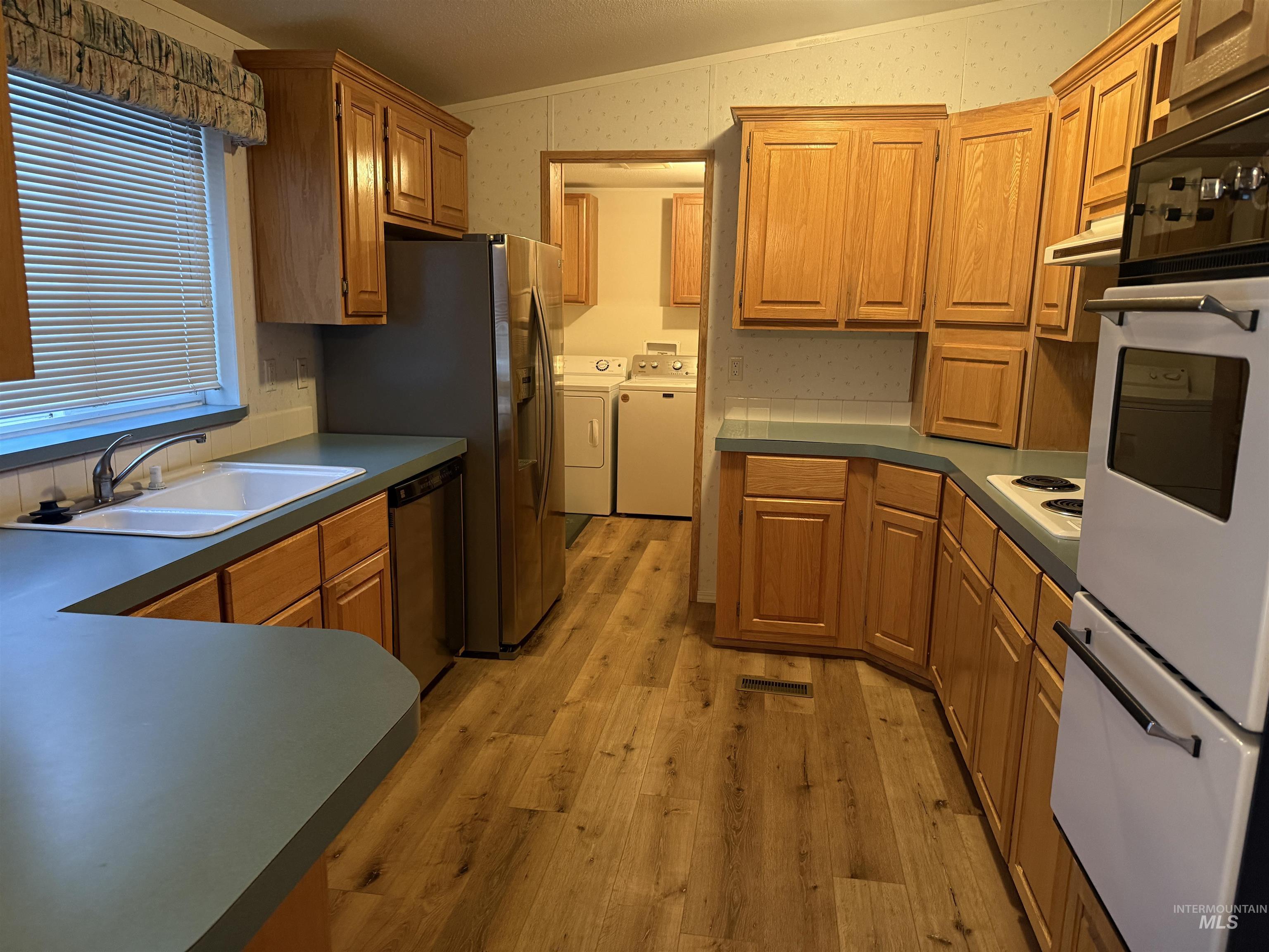 Kitchen with appliances with stainless steel finishes, light wood-style floors, wallpapered walls, washing machine and dryer, and brown cabinetry
