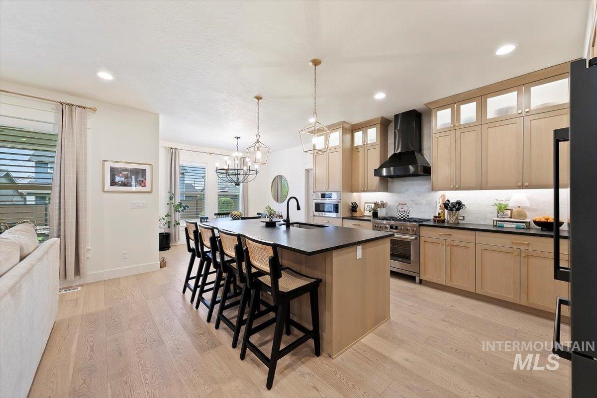 Kitchen featuring backsplash, dark countertops, an island with sink, wall chimney exhaust hood, and a kitchen breakfast bar
