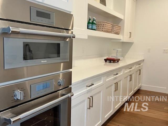 Kitchen with stainless steel double oven, white cabinetry, dark wood finished floors, open shelves, and light stone countertops