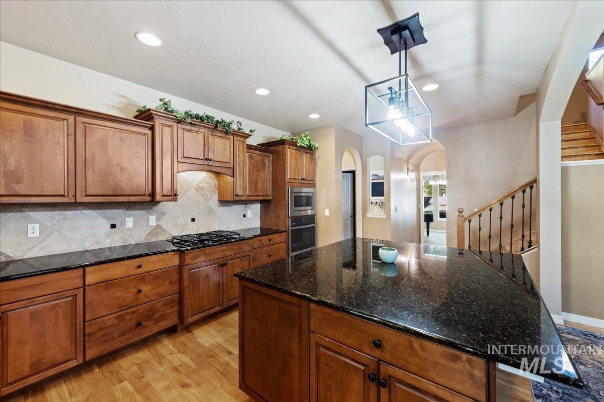 Kitchen featuring brown cabinets, dark stone countertops, light wood-style floors, tasteful backsplash, and hanging light fixtures