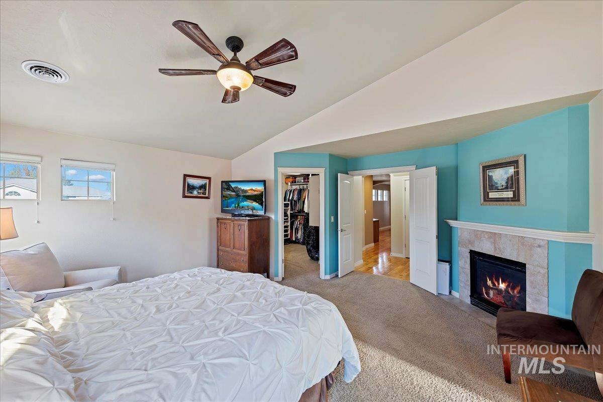 Carpeted bedroom with vaulted ceiling, a walk in closet, a tiled fireplace, and ceiling fan