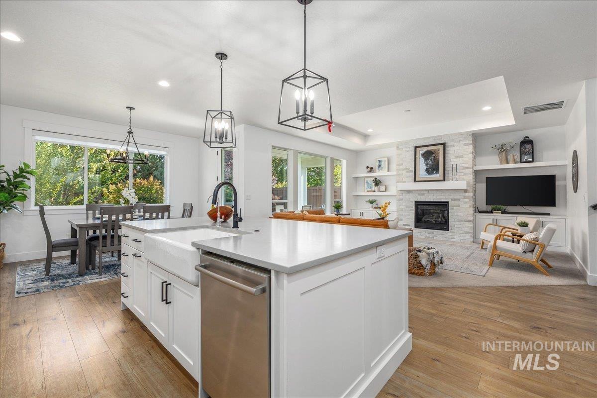 Kitchen featuring white cabinets, a raised ceiling, a stone fireplace, a kitchen island with sink, and decorative light fixtures