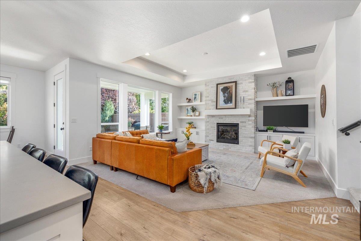 Living area with a raised ceiling, light wood-type flooring, a stone fireplace, and recessed lighting