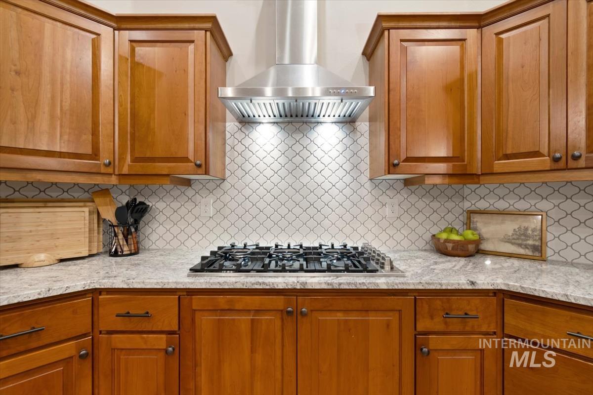 Kitchen featuring wall chimney range hood, brown cabinetry, decorative backsplash, light stone counters, and stainless steel gas cooktop