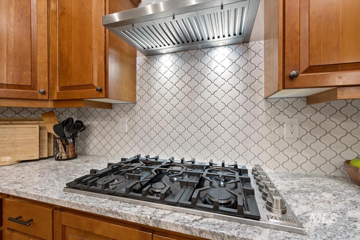 Kitchen with wall chimney exhaust hood, decorative backsplash, stainless steel gas cooktop, brown cabinets, and light stone countertops