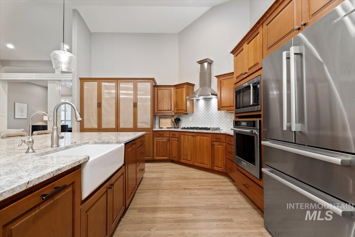 Kitchen featuring appliances with stainless steel finishes, brown cabinetry, backsplash, light wood finished floors, and wall chimney range hood