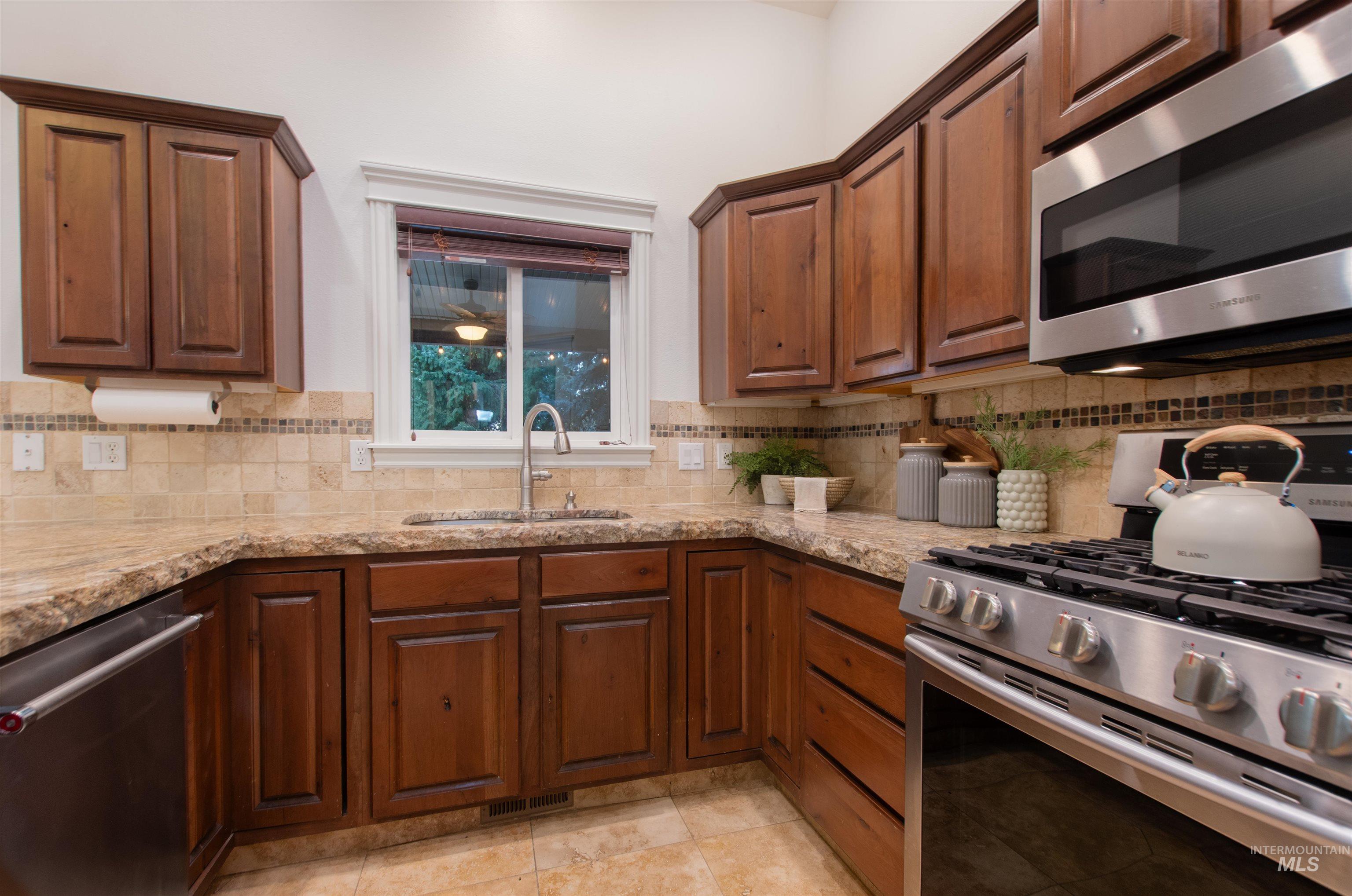 Kitchen featuring stainless steel appliances, decorative backsplash, light stone countertops, brown cabinetry, and light tile patterned floors