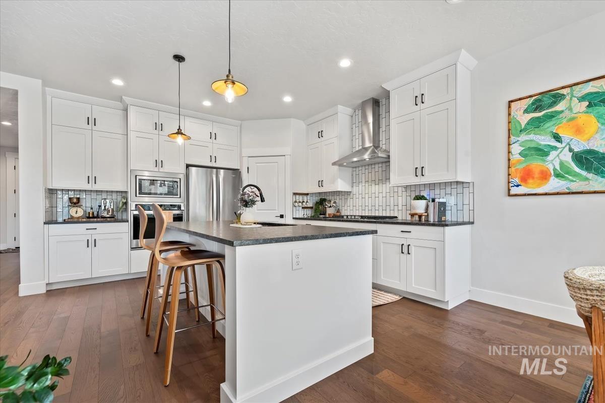 Kitchen featuring white cabinets, stainless steel appliances, an island with sink, wall chimney range hood, and a kitchen breakfast bar