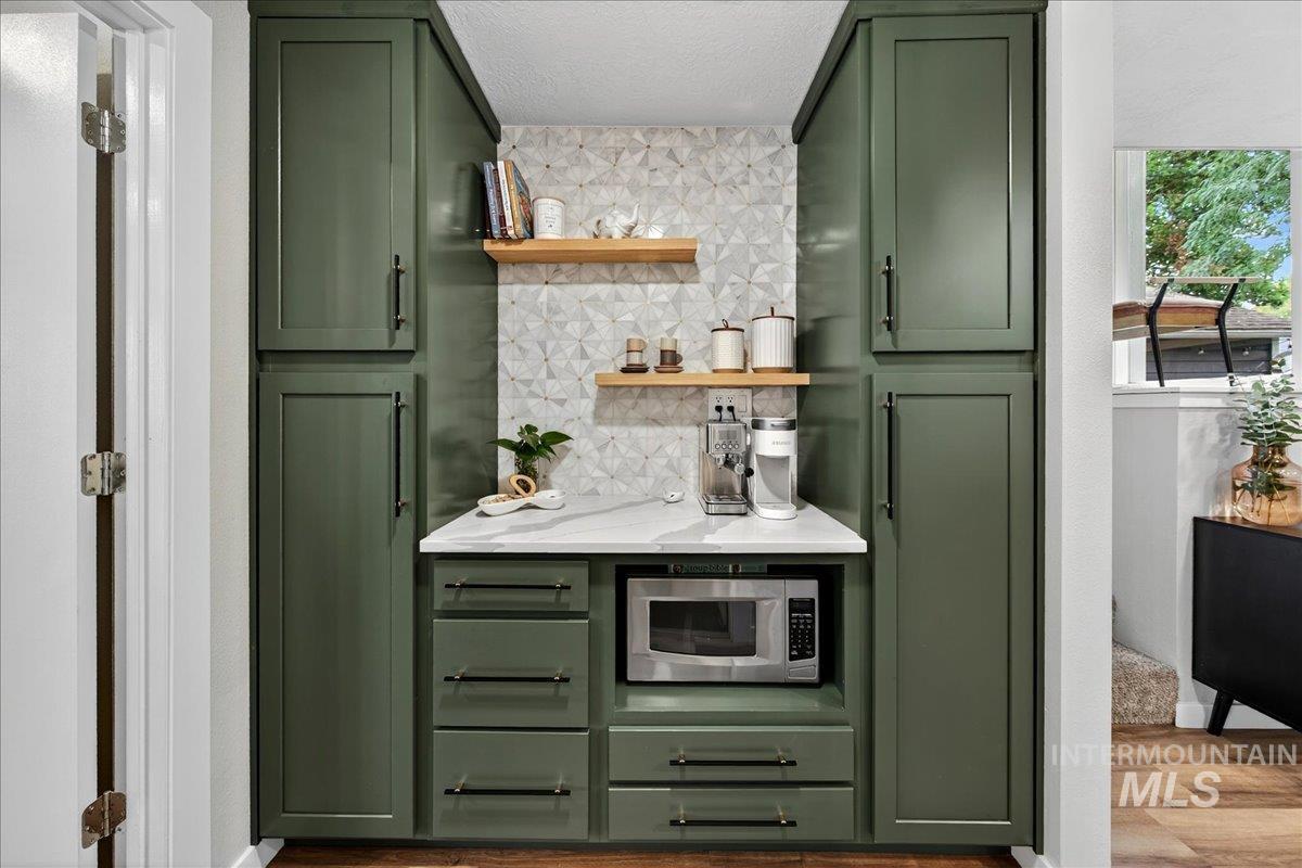 Bar area with green cabinetry, backsplash, wood finished floors, open shelves, and stainless steel microwave
