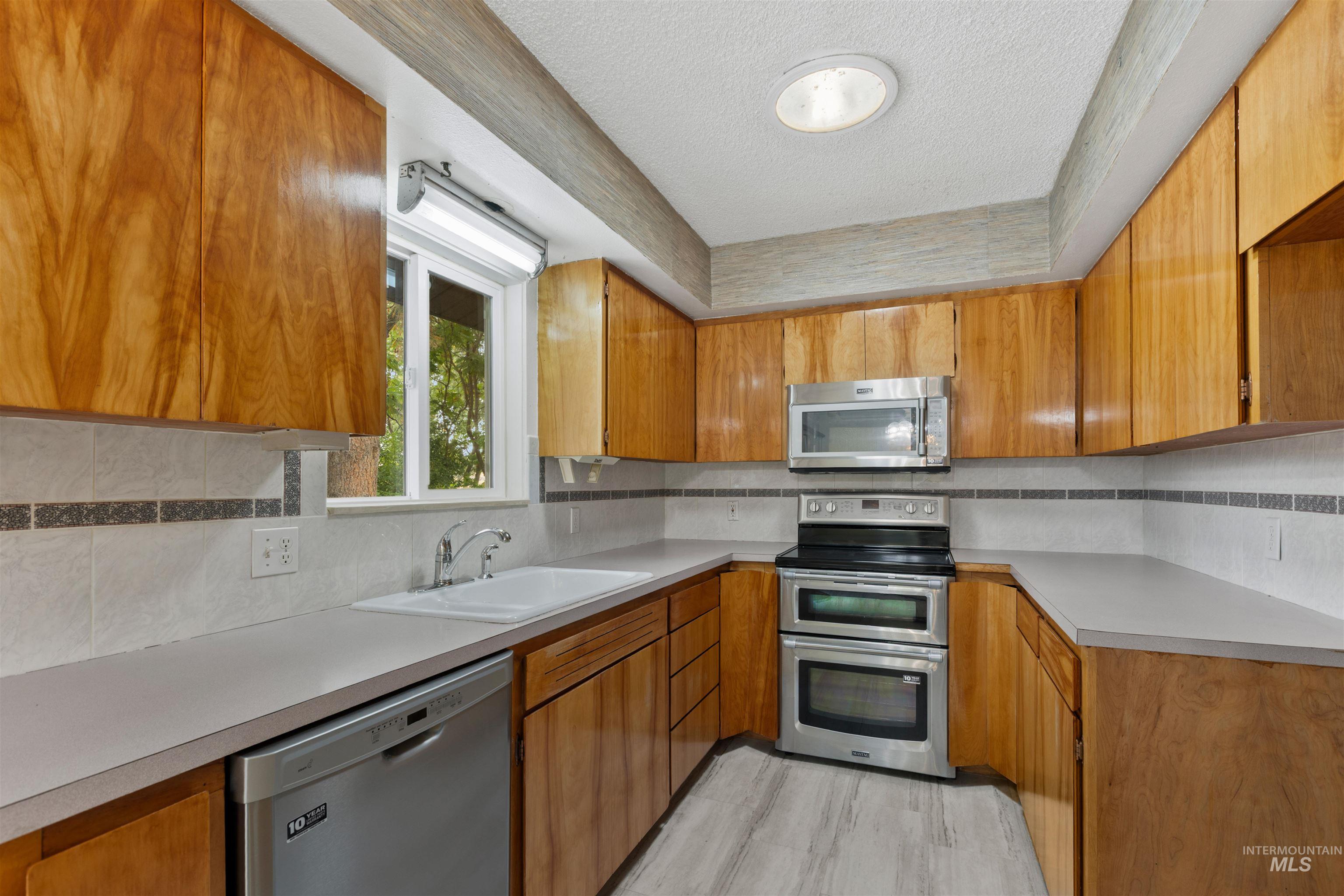 Kitchen with stainless steel appliances, tasteful backsplash, brown cabinetry, light countertops, and a textured ceiling