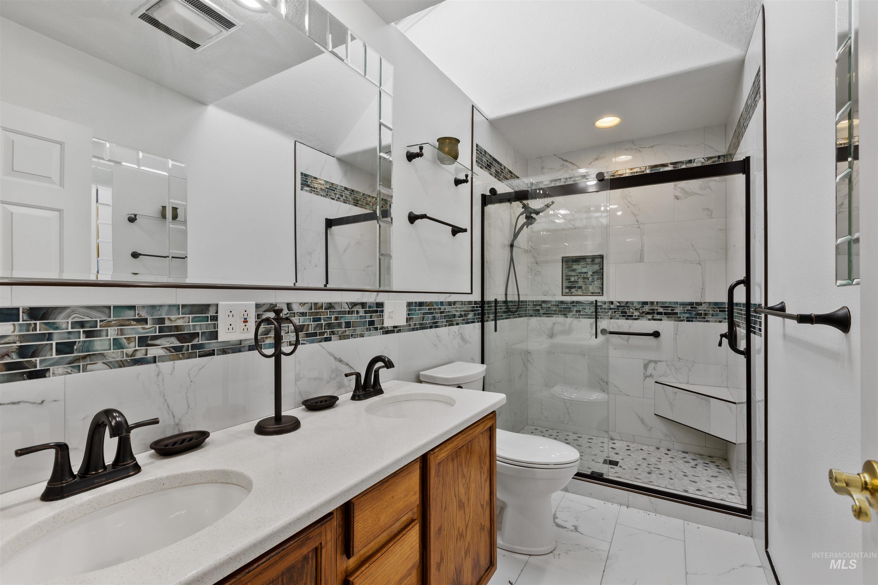 Full bath with double vanity, a marble finish shower, light marble finish flooring, tile walls, and backsplash
