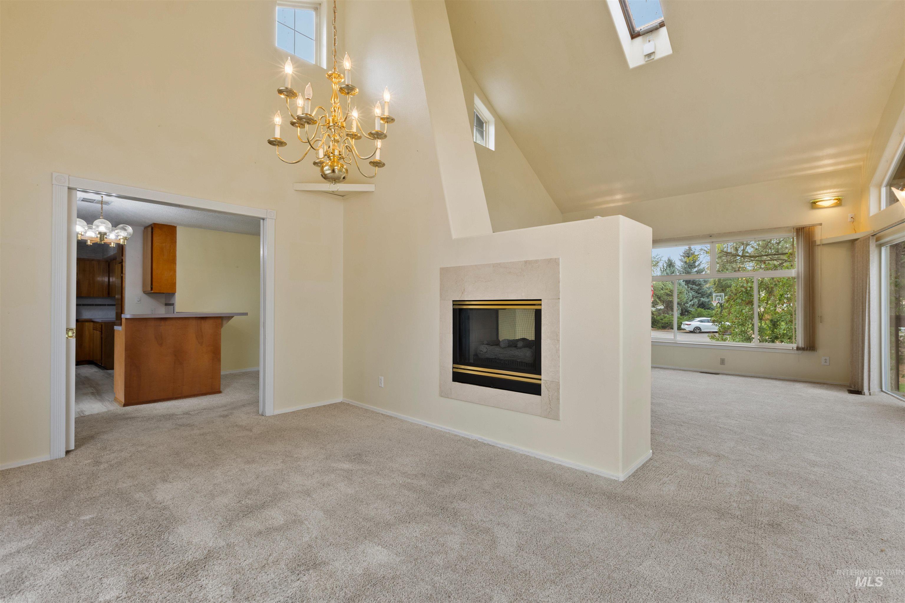 Unfurnished living room with high vaulted ceiling, light colored carpet, a multi sided fireplace, a chandelier, and a skylight