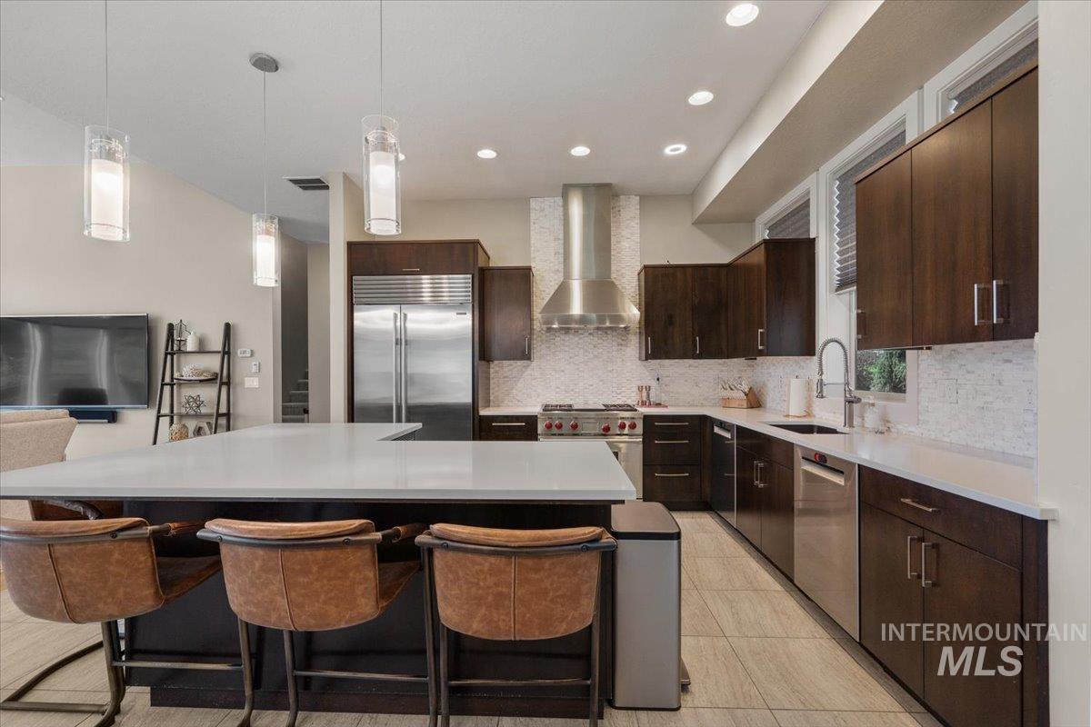 Kitchen featuring high quality appliances, wall chimney exhaust hood, backsplash, dark brown cabinets, and a kitchen island