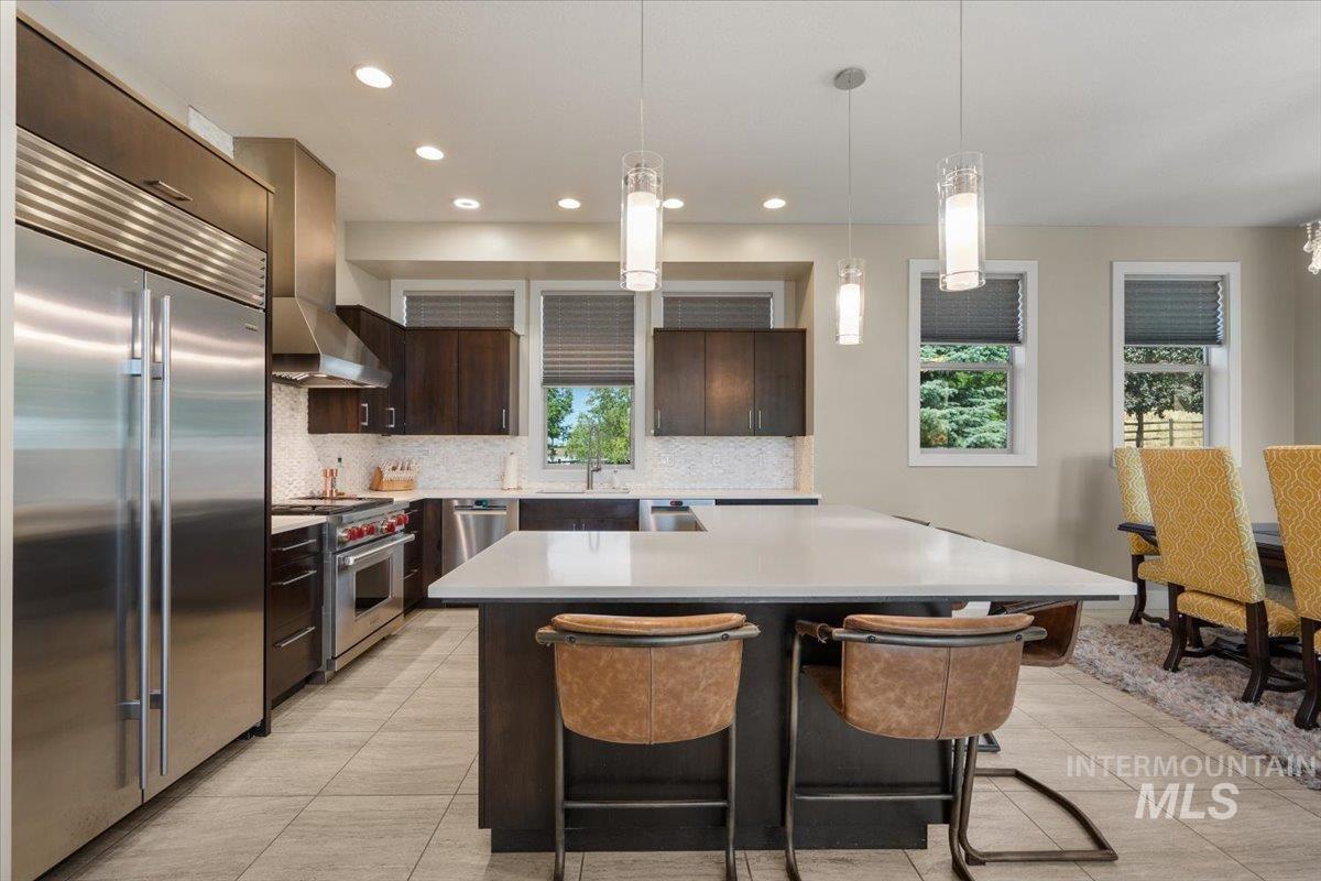 Kitchen featuring high quality appliances, light countertops, wall chimney exhaust hood, backsplash, and dark brown cabinets