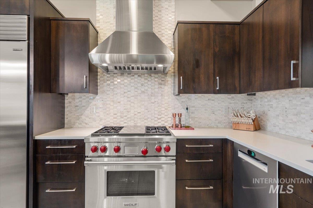 Kitchen featuring wall chimney exhaust hood, high end appliances, dark brown cabinets, and decorative backsplash