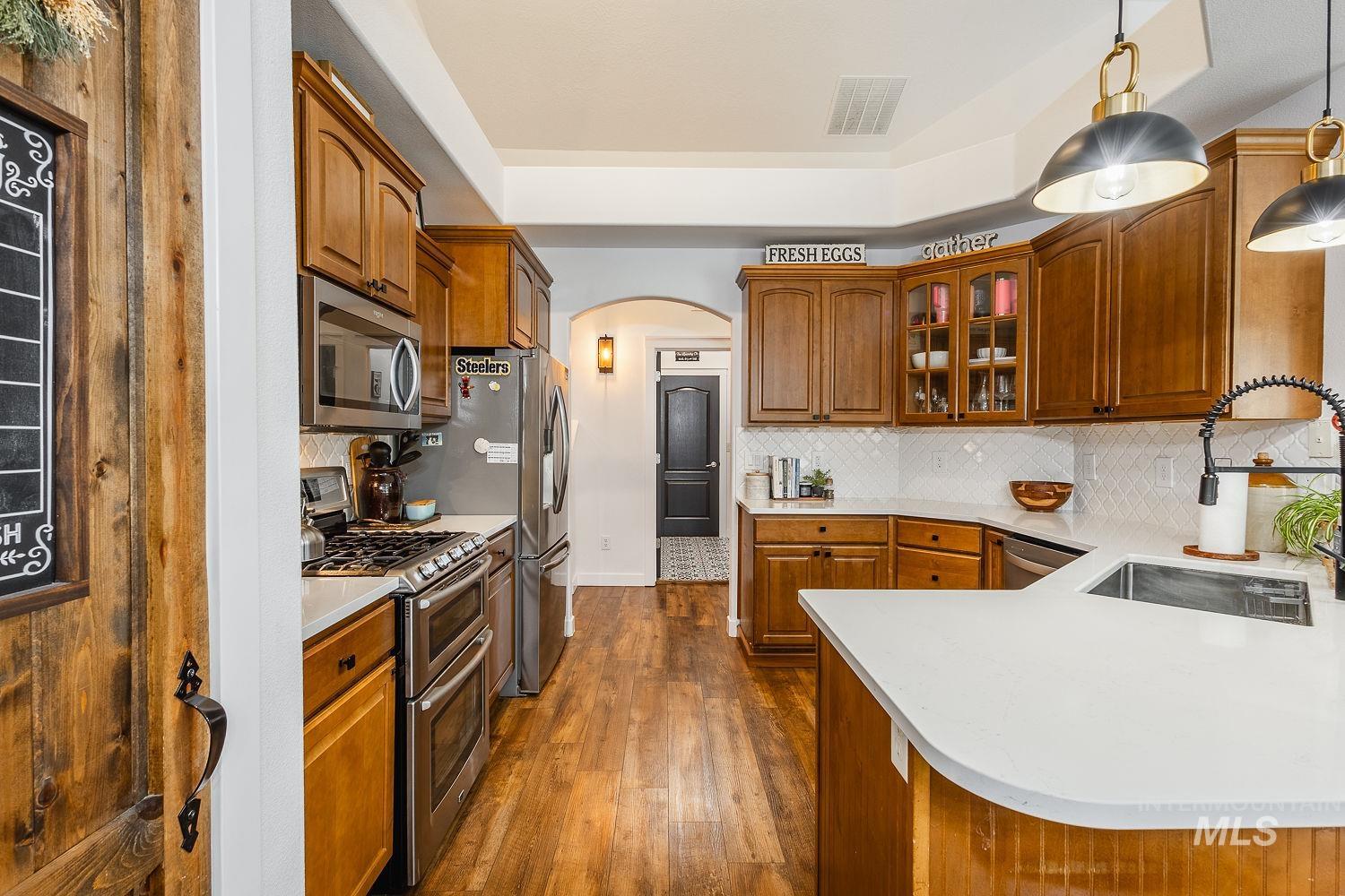 Kitchen with arched walkways, appliances with stainless steel finishes, glass insert cabinets, brown cabinets, and dark wood-type flooring