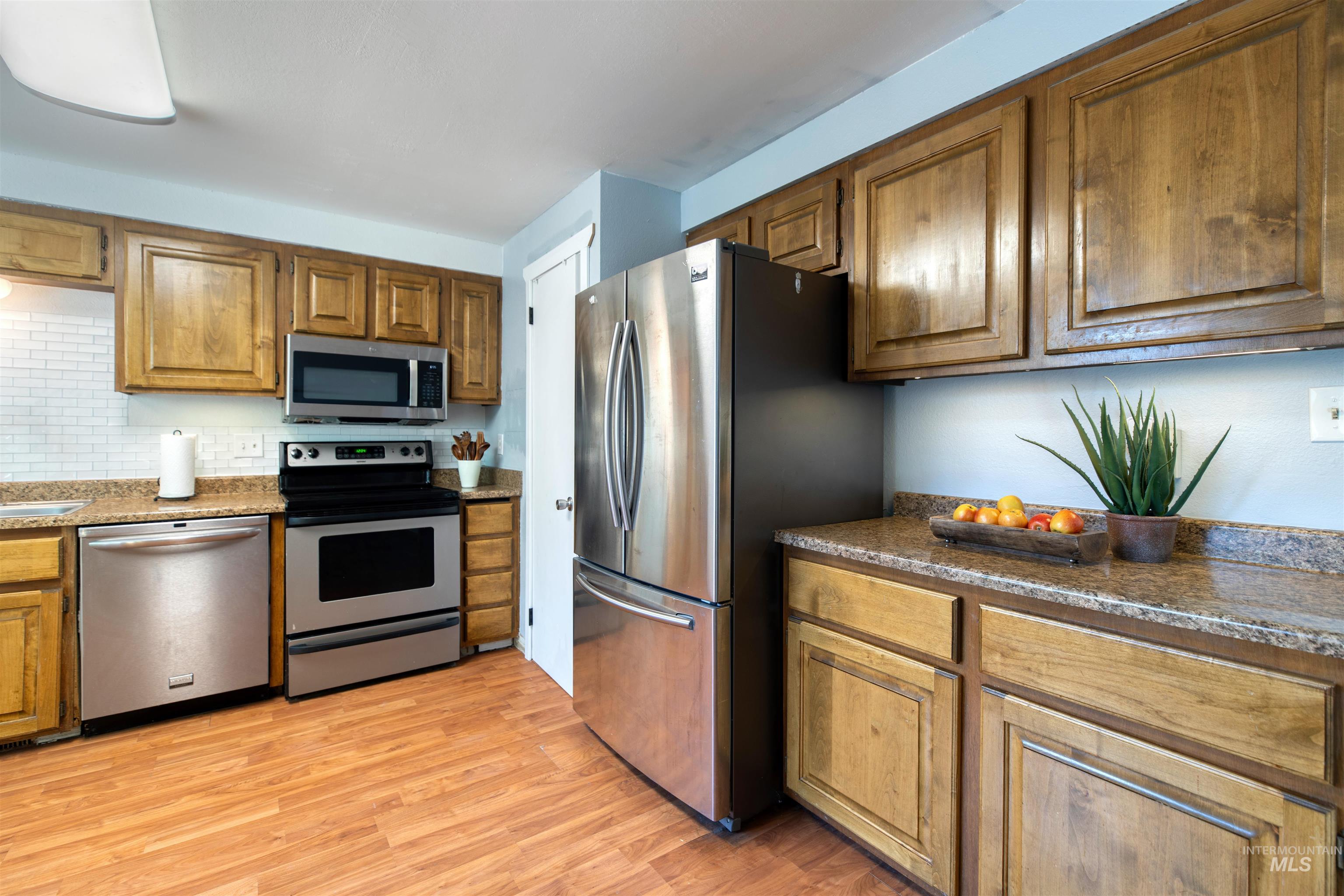 Kitchen featuring stainless steel appliances, brown cabinetry, light wood-type flooring, backsplash, and dark stone countertops