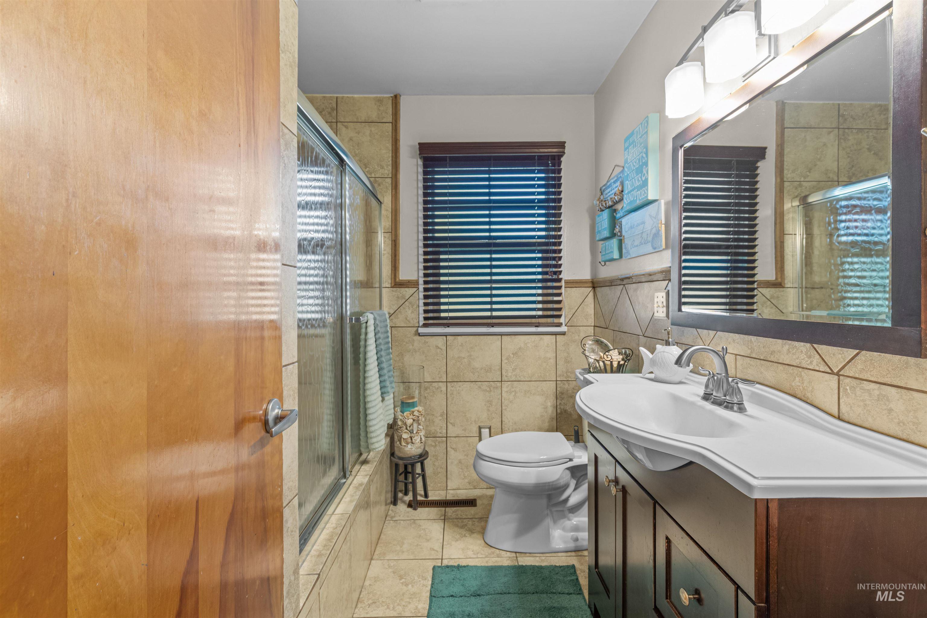 Full bath with light tile patterned floors, vanity, tile walls, shower / bath combination with glass door, and a wainscoted wall