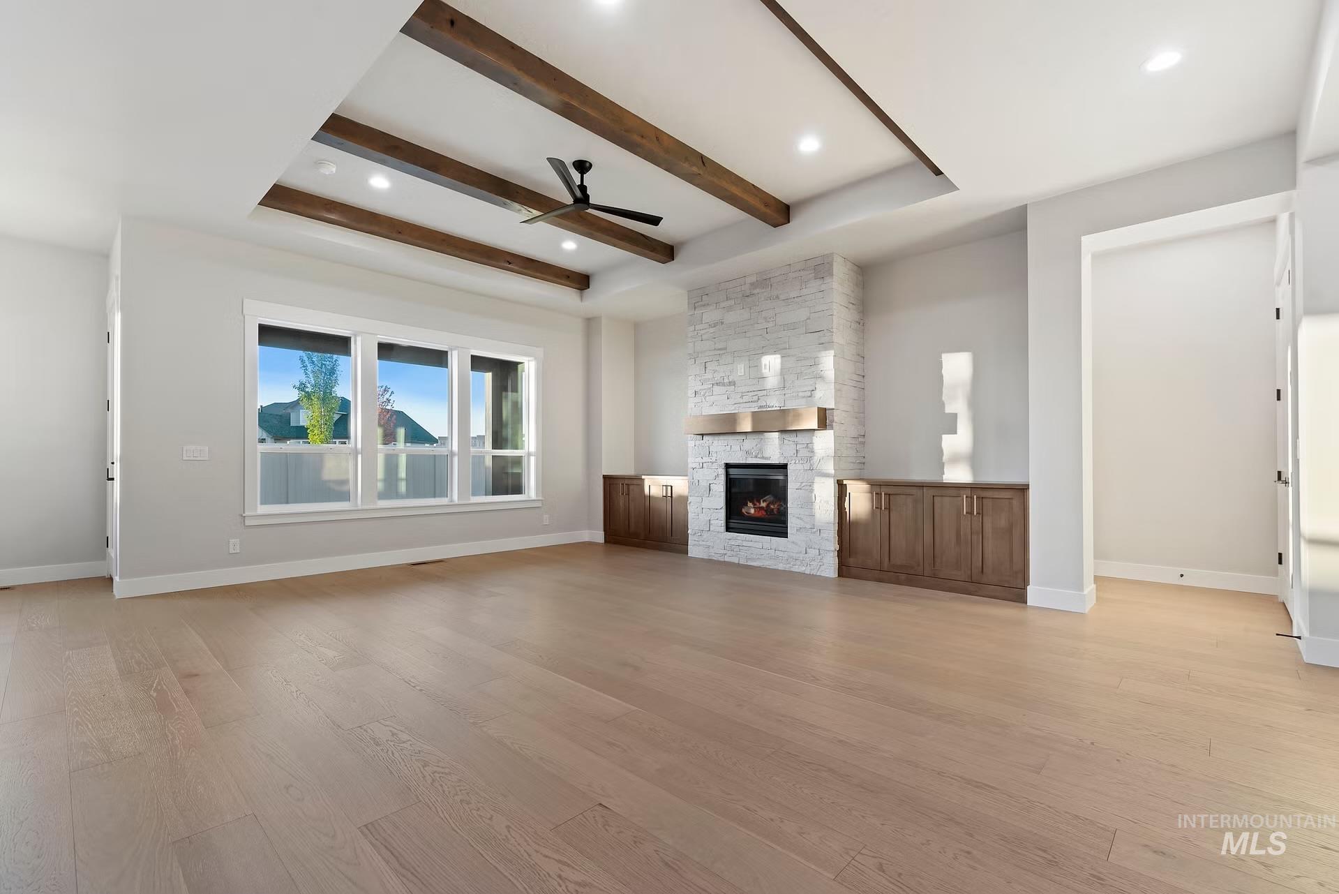 Unfurnished living room with ceiling fan, beam ceiling, light wood-style flooring, a stone fireplace, and a tray ceiling
