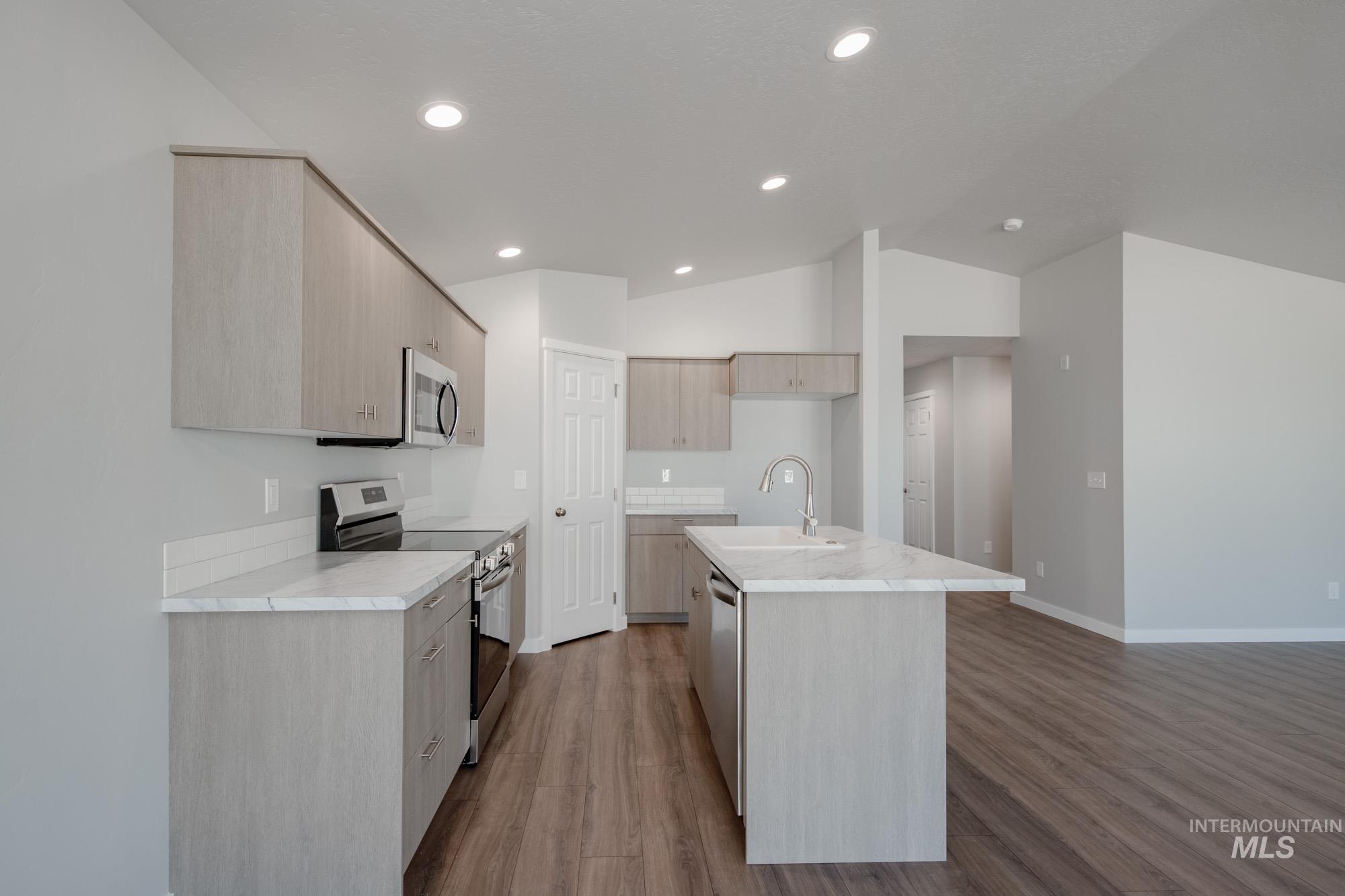 Kitchen with appliances with stainless steel finishes, light wood finished floors, vaulted ceiling, light brown cabinetry, and an island with sink