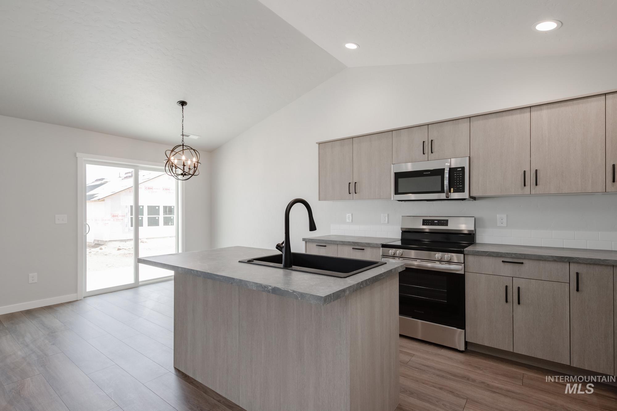 Kitchen featuring light brown cabinets, stainless steel appliances, a center island with sink, vaulted ceiling, and light wood-type flooring