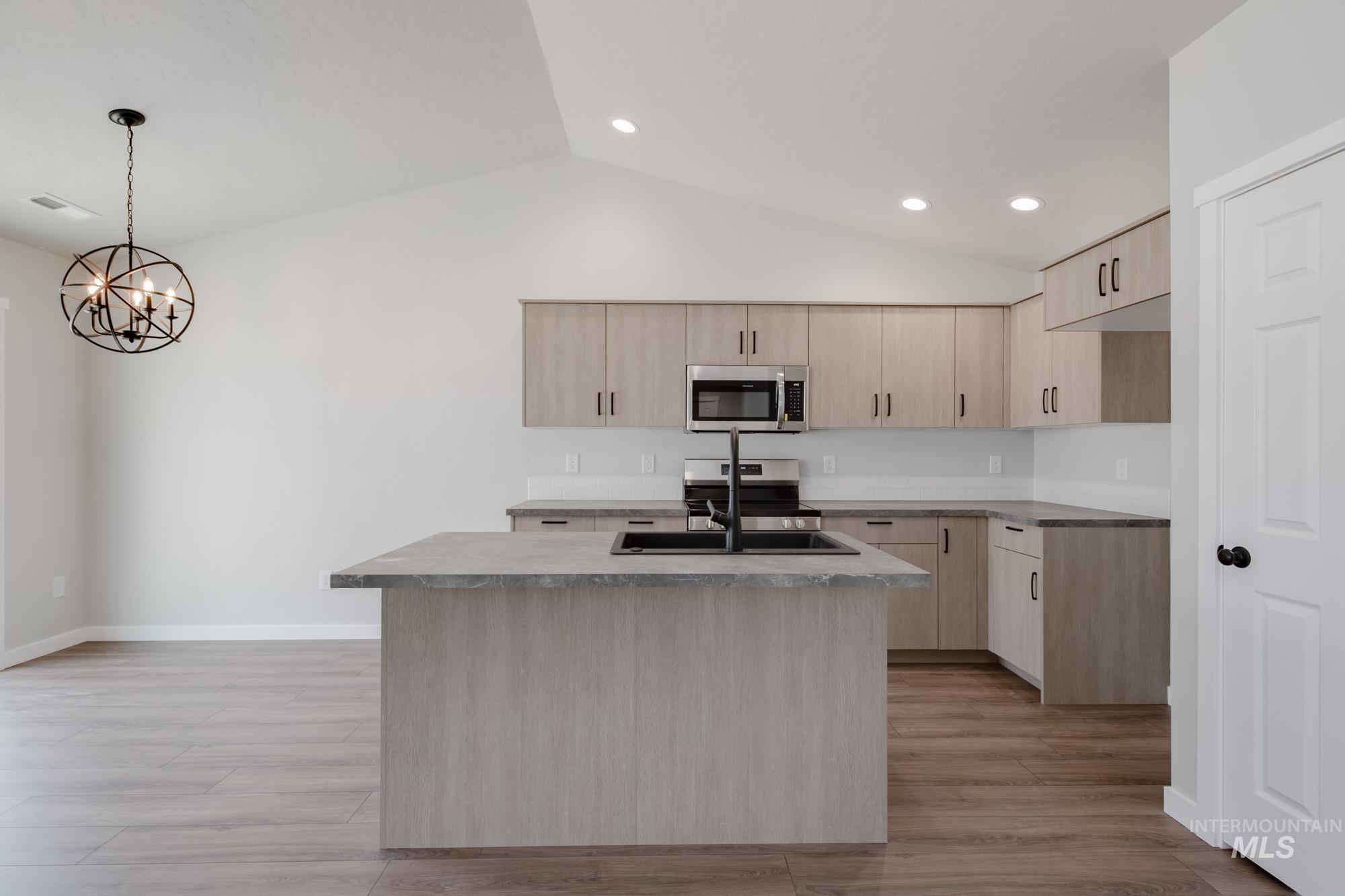 Kitchen with light brown cabinetry, stainless steel appliances, vaulted ceiling, a center island with sink, and light wood-style floors