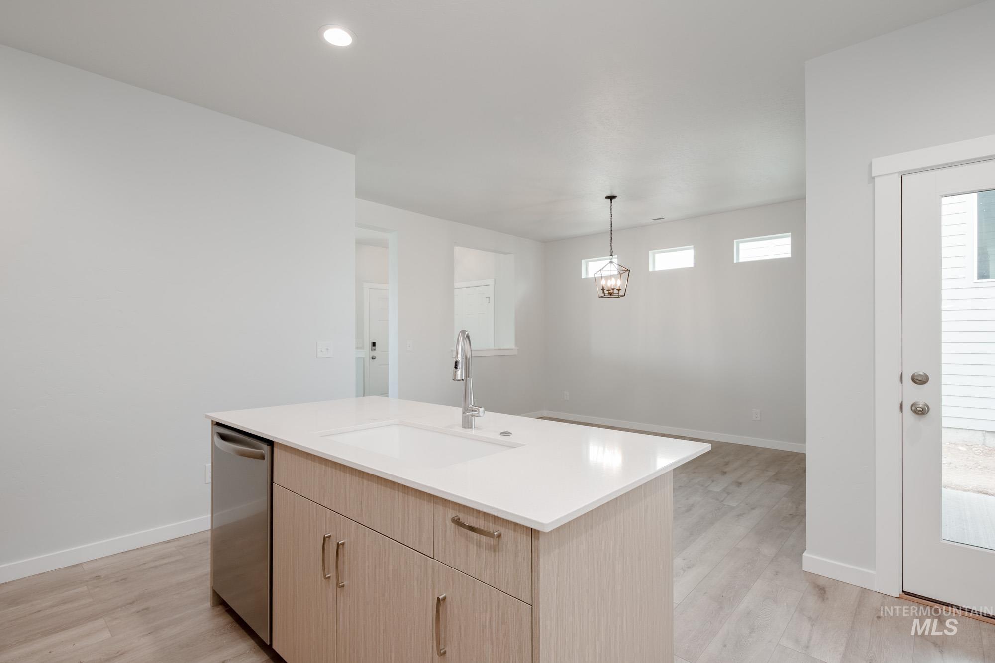 Kitchen with light brown cabinetry, light wood-style floors, dishwasher, hanging light fixtures, and a center island with sink