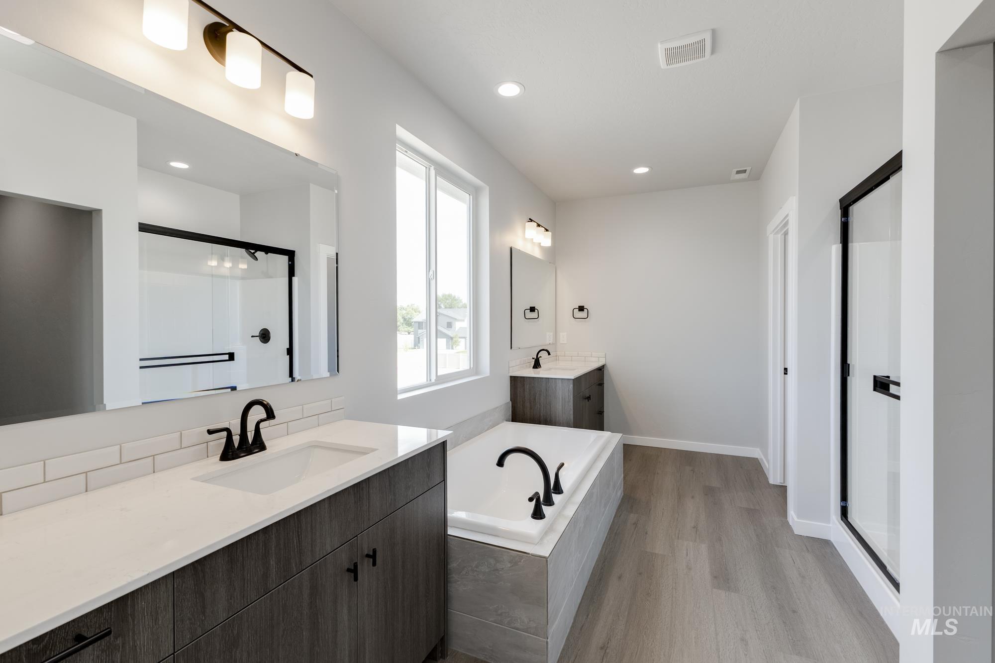 Full bath with two vanities, a shower stall, a garden tub, light wood-style flooring, and recessed lighting