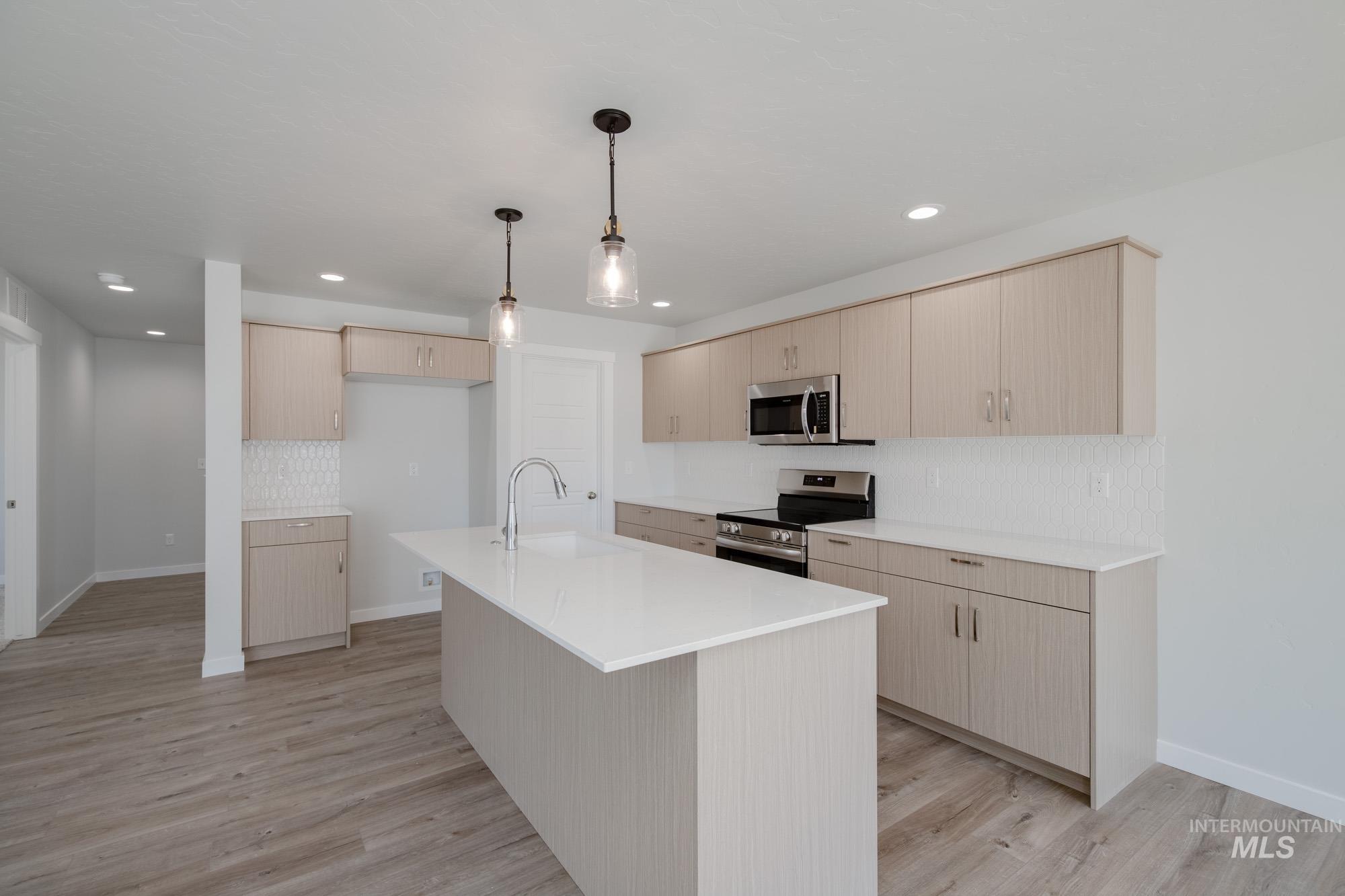Kitchen featuring light brown cabinets, appliances with stainless steel finishes, decorative backsplash, hanging light fixtures, and light wood-style flooring