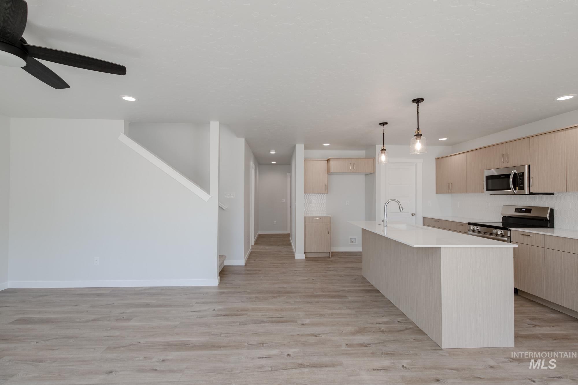 Kitchen with light brown cabinets, light wood-style floors, appliances with stainless steel finishes, hanging light fixtures, and a kitchen island with sink