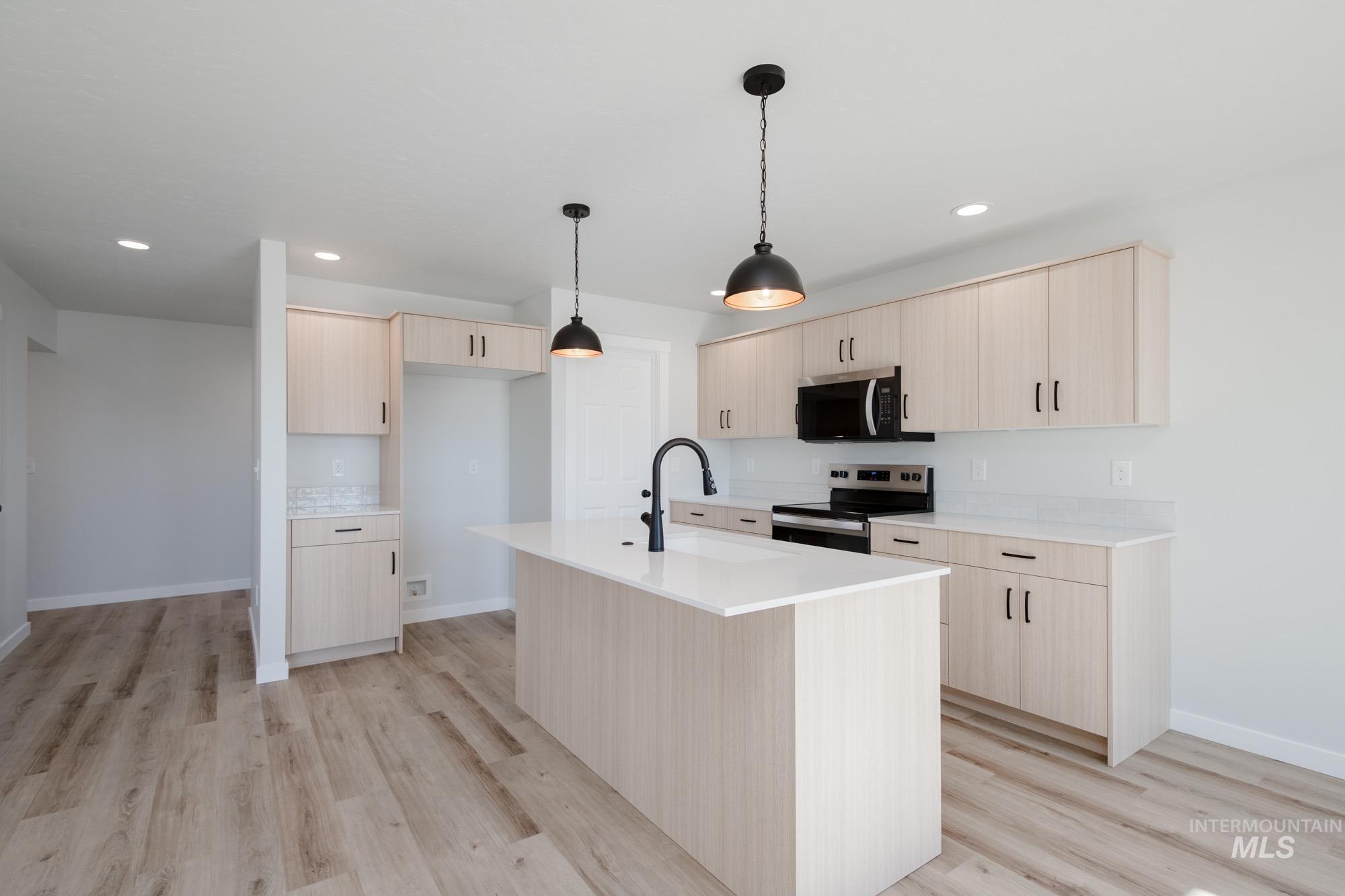 Kitchen with light brown cabinetry, stainless steel appliances, light wood-style flooring, decorative light fixtures, and an island with sink